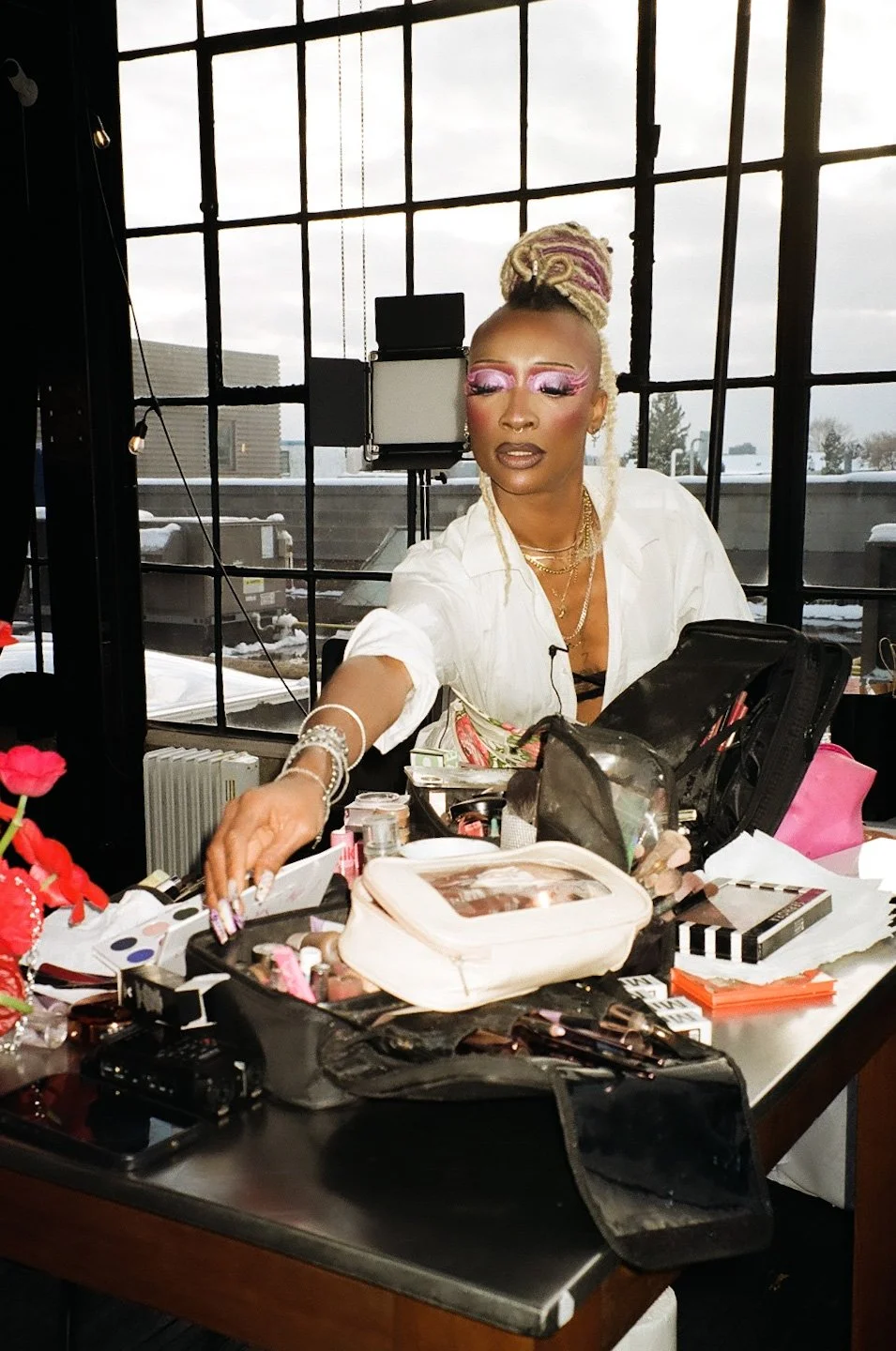 A woman with elaborate makeup and jewelry sits at a cluttered makeup station with various cosmetics and tools, in front of a large window showing an overcast sky.