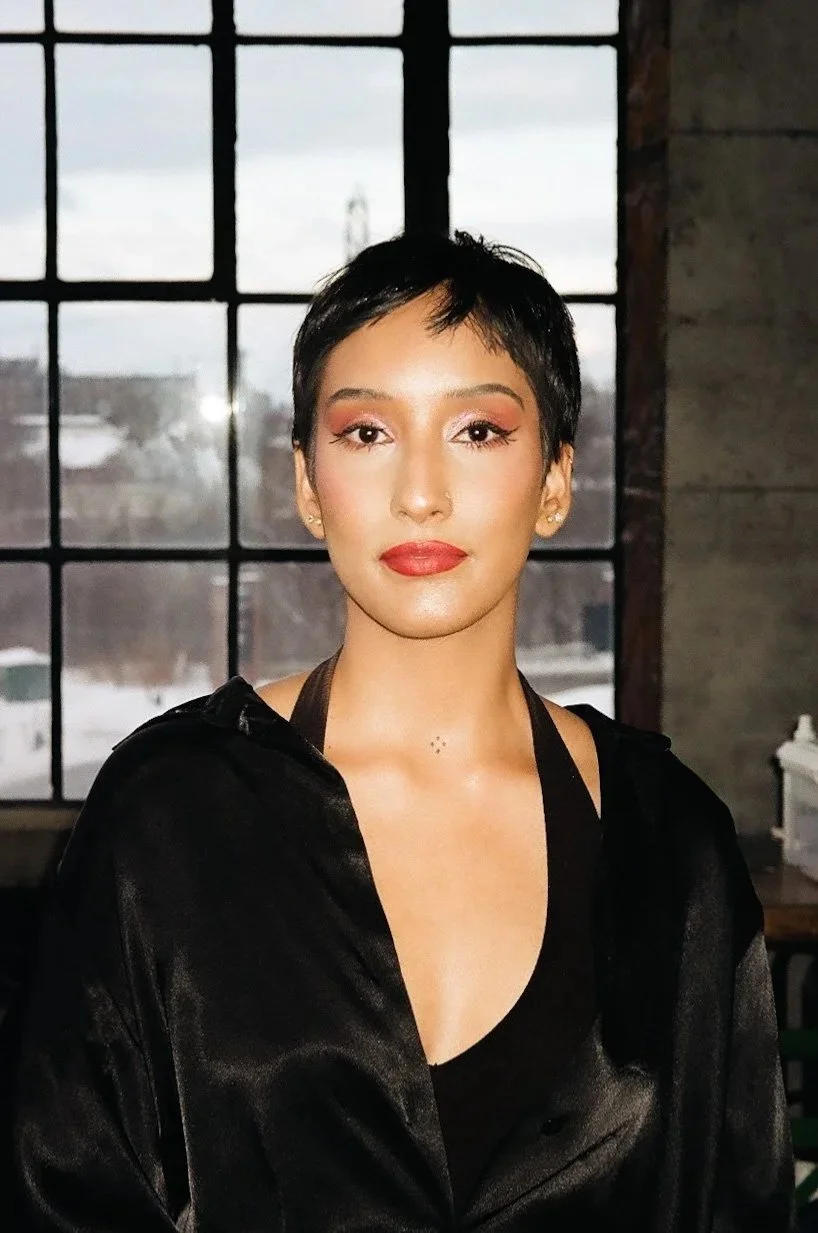 A woman with short black hair and makeup, wearing a black satin jacket, standing indoors near a large window with a snowy outdoor scene visible behind her.
