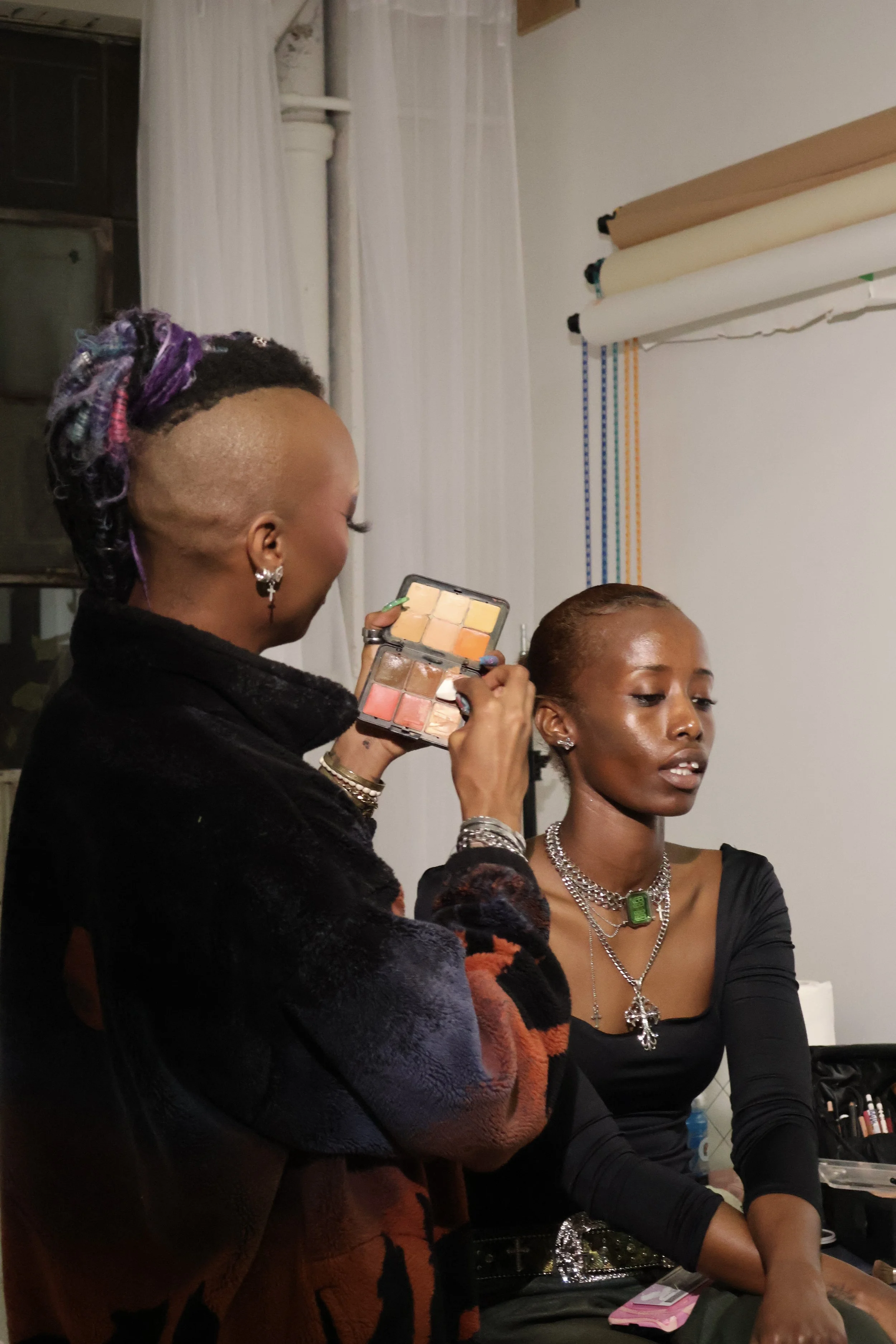 A woman applying makeup to a young woman seated in front of her in a studio setting.