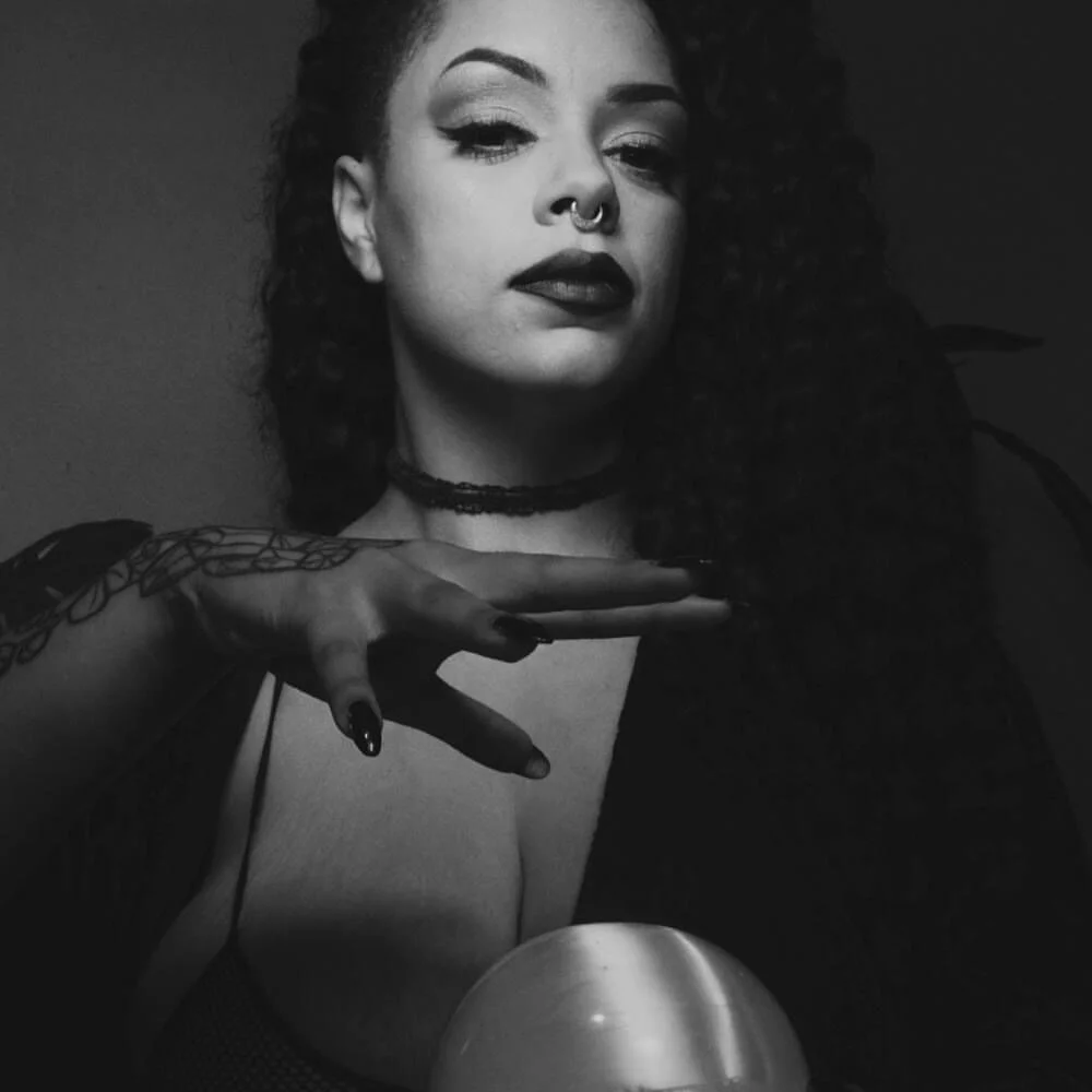 A woman with curly hair and bold makeup, wearing dark lipstick and a septum ring, poses with her hand near her face in a black and white photo.