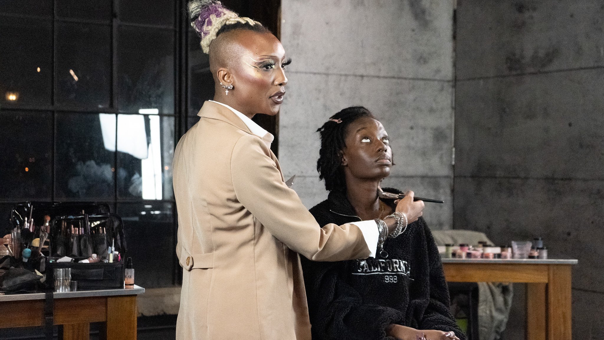 A makeup artist applying makeup to a woman seated in a salon or studio space with gray concrete walls and tools on tables.