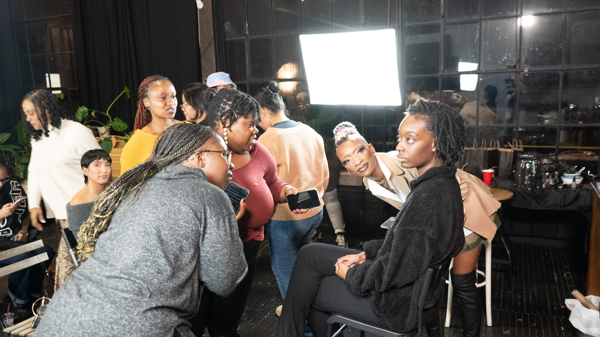 Group of women in a discussion at a social gathering or event, with a woman sitting on a chair and others standing around her.