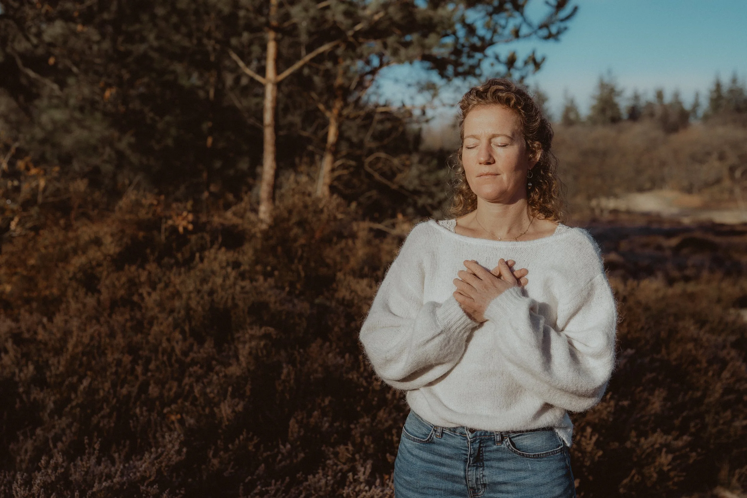Vrouw met ogen dicht en handen op haar borst staand in de natuur, aardend, zacht, in ochtendlicht.