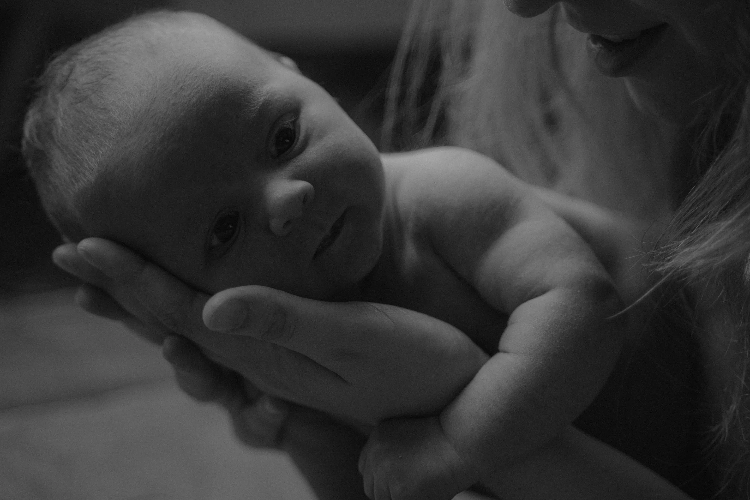 Zwart wit foto van een kleine baby in de handen van mama. Newborn fotoshoot thuis, Newborn fotografie, tijdloze fotografie. Assen Drenthe, Noord-Nederland. Footprint Stories Fotografie, verhalende fotografie. Documentaire Newborn reportage thuis.