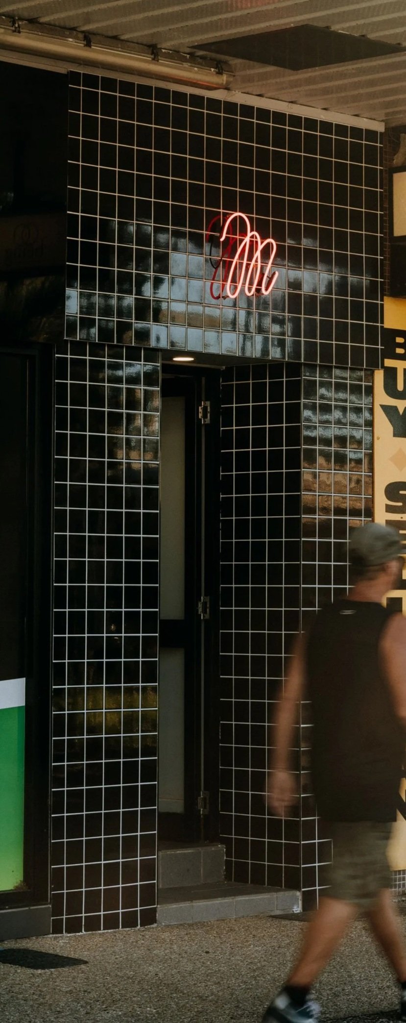 Pedestrian walking past a black tiled storefront with a neon red sign and a partially closed shutter, with advertising signs above.