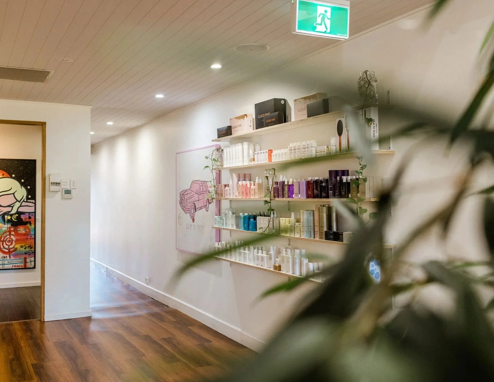A boutique shop interior with shelves filled with colorful bottles, a wall with a pink car illustration and whiteboard, and a doorway showing a colorful mural featuring a sleeping figure.