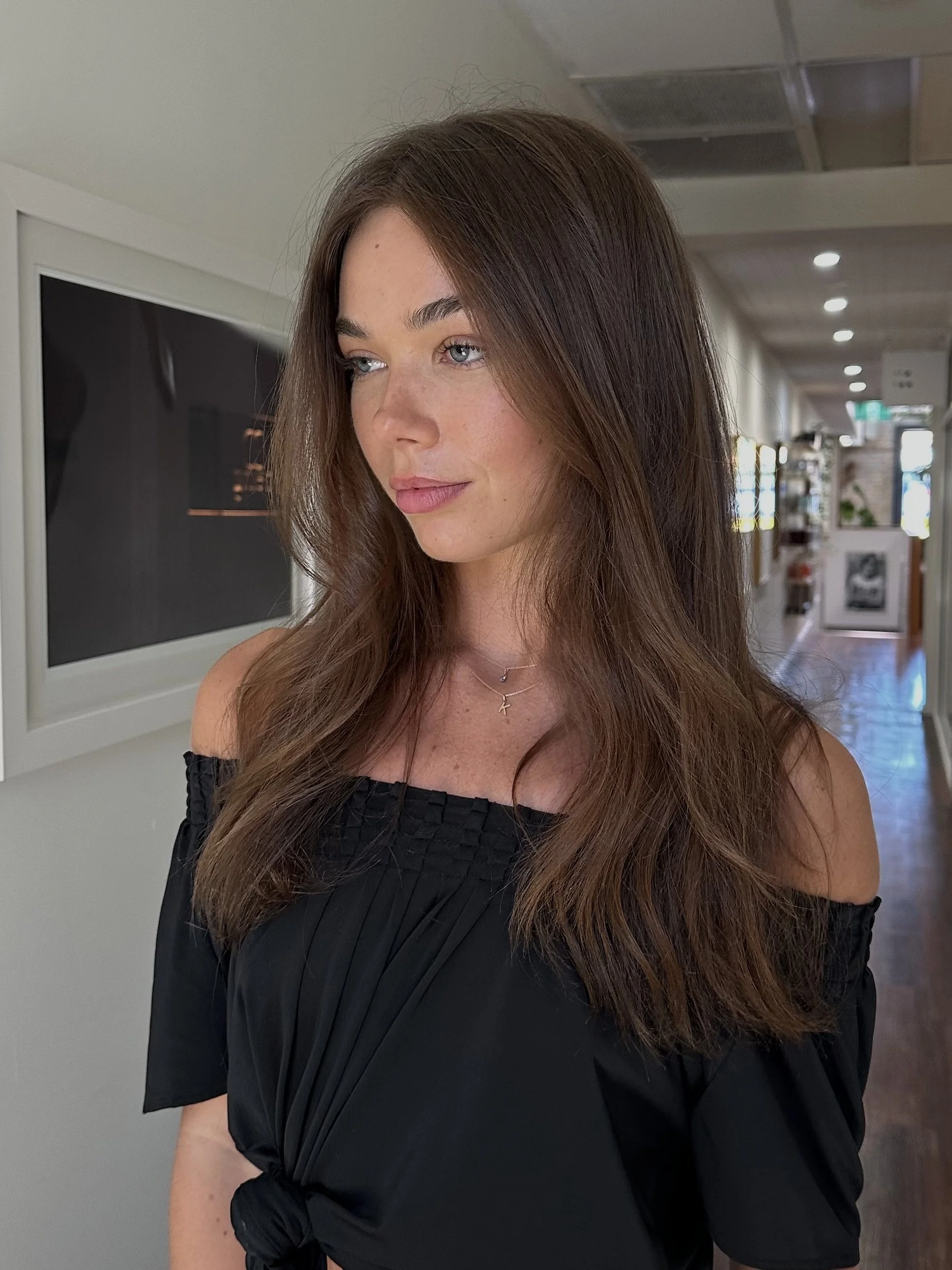 A woman with long brown hair wearing a black off-the-shoulder top and layered necklaces, standing indoors with a hallway and artwork in the background.