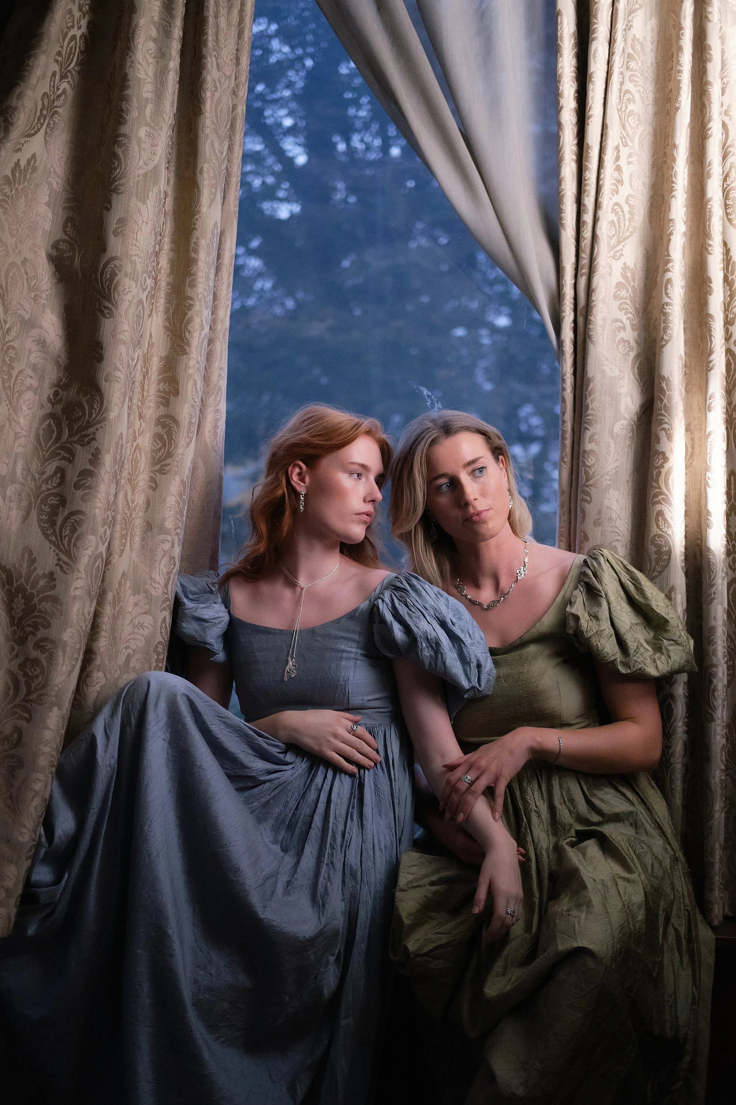 Two women dressed in period costumes sitting together by a window with gold and cream patterned curtains, looking thoughtfully outside as evening falls.
