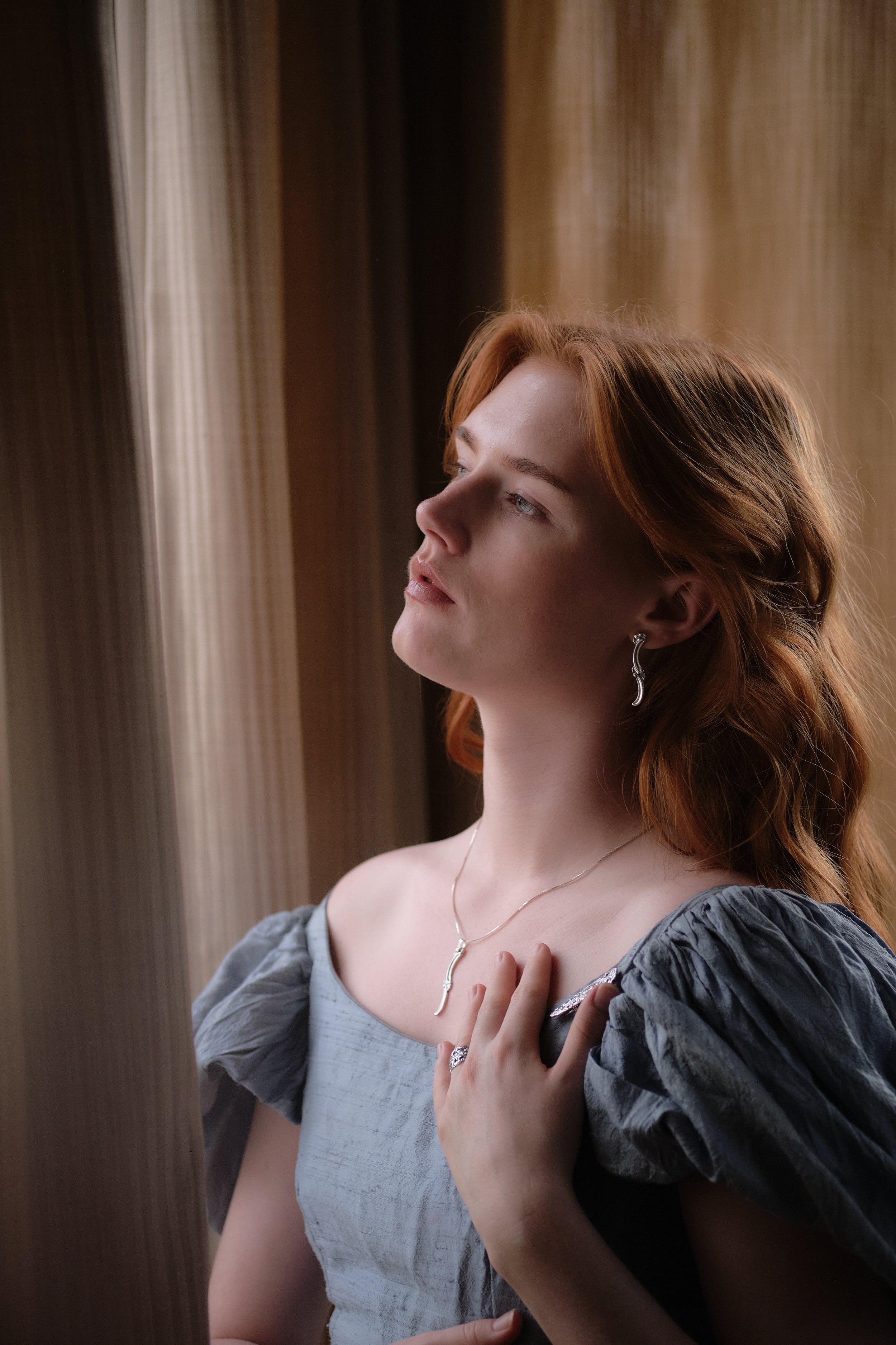 A woman with red hair and light skin gazes thoughtfully out a window, touching her chest with one hand, dressed in a gray puff-sleeve top and silver jewelry.