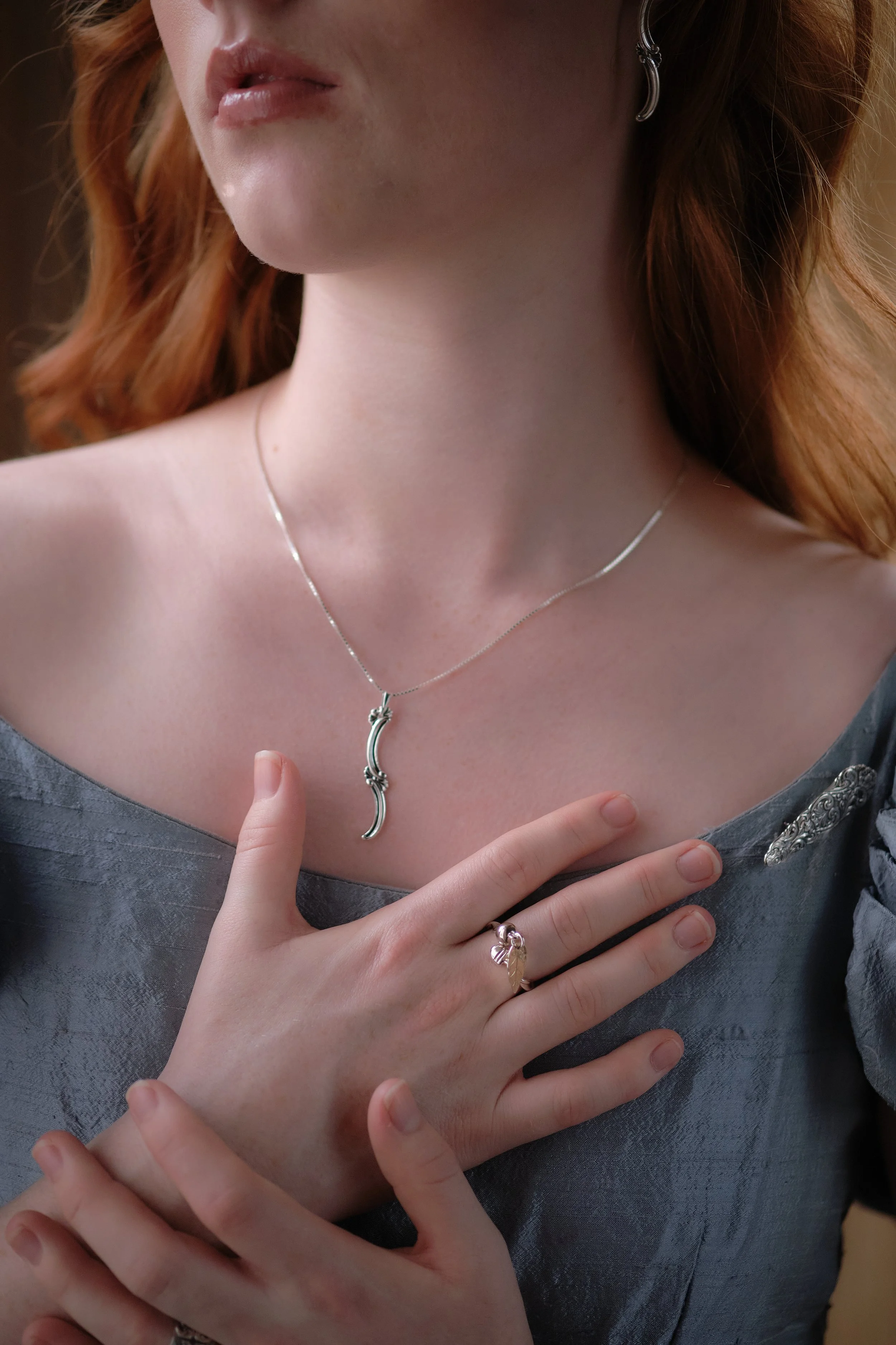 Close-up of a woman with red hair wearing a silver necklace with a unique curved pendant and a silver ring with a pink gemstone on her finger. She is touching her chest with her right hand, dressed in a gray satin-like dress with decorative details.