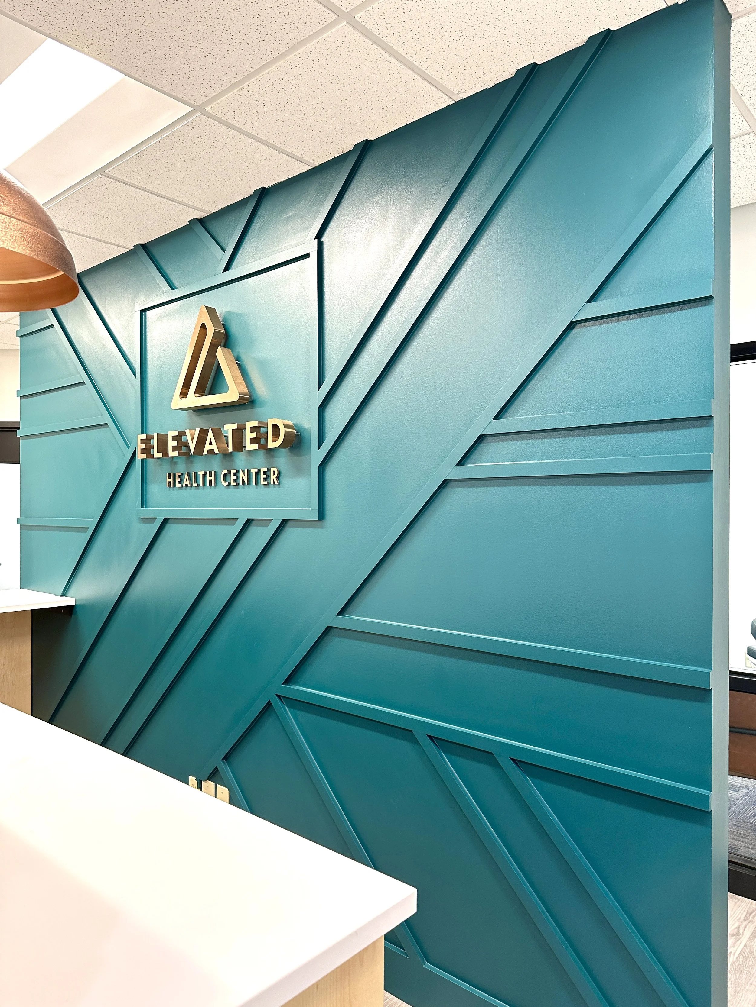 Office reception area with a teal geometric accent wall displaying a logo sign for Elevated Health Center.