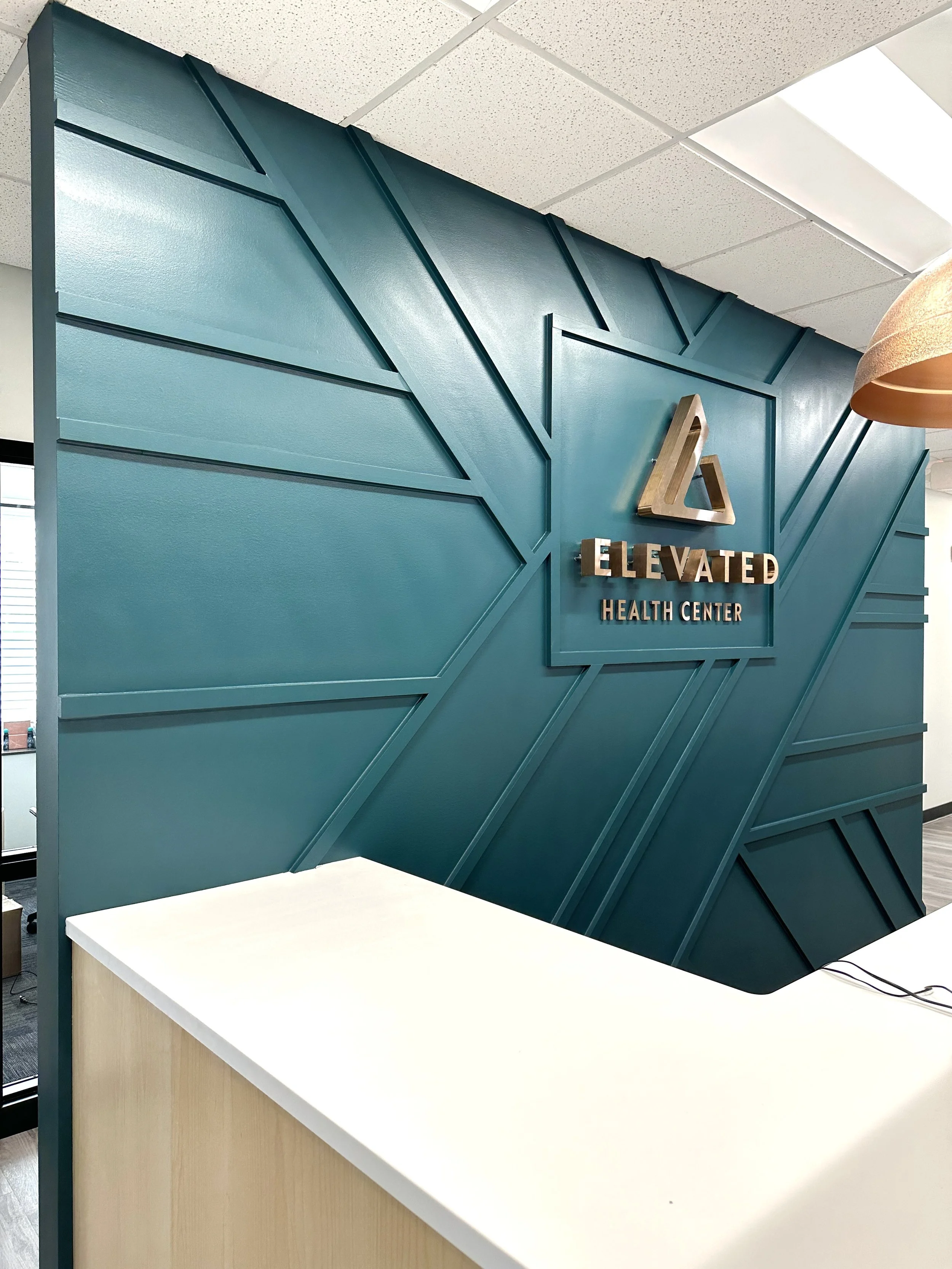 Office reception area with a teal geometric accent wall displaying a logo sign for Elevated Health Center.