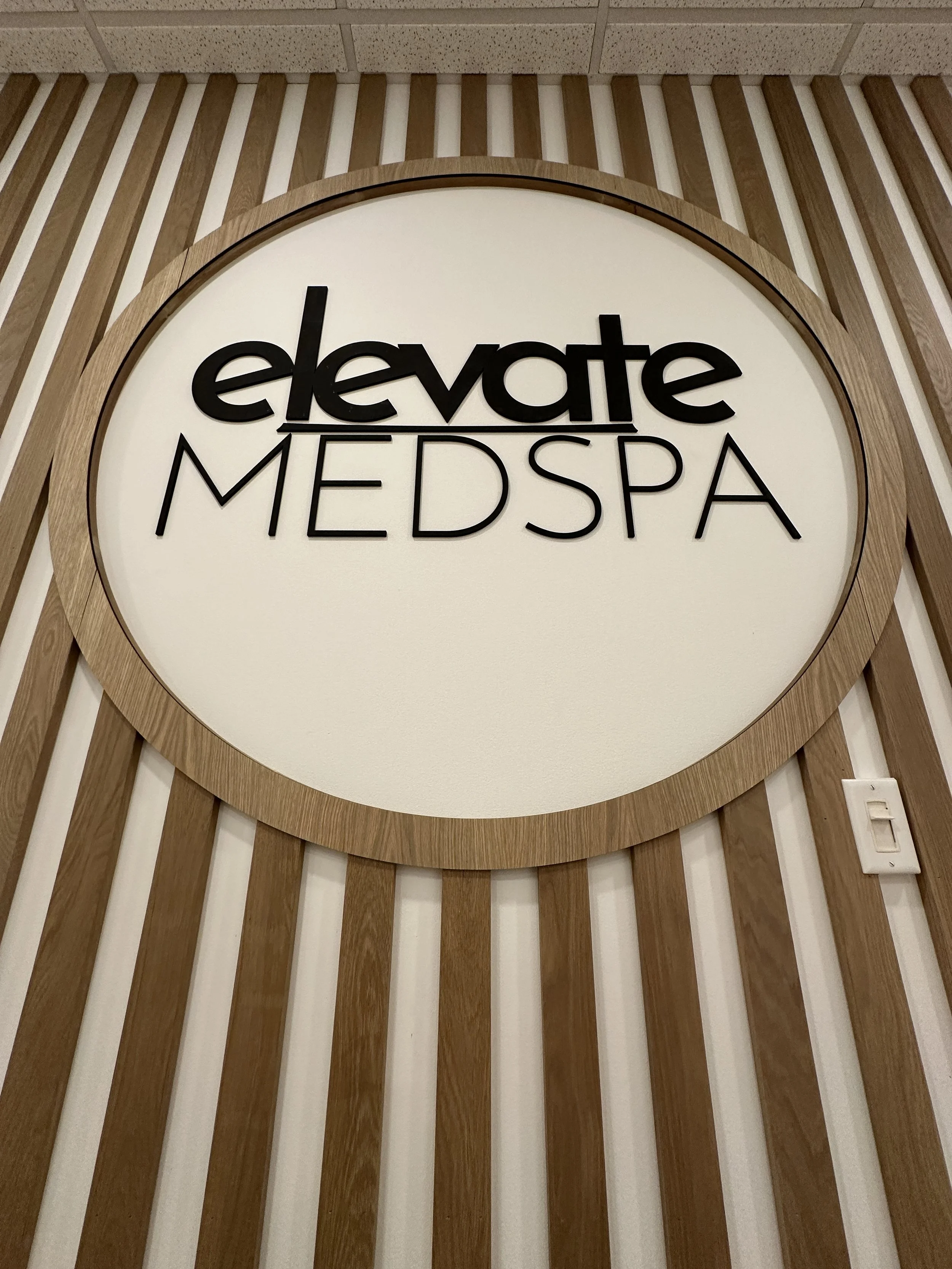 A round sign mounted on a wood-paneled wall, displaying the words 'elevate MEDSPA' in black lettering.