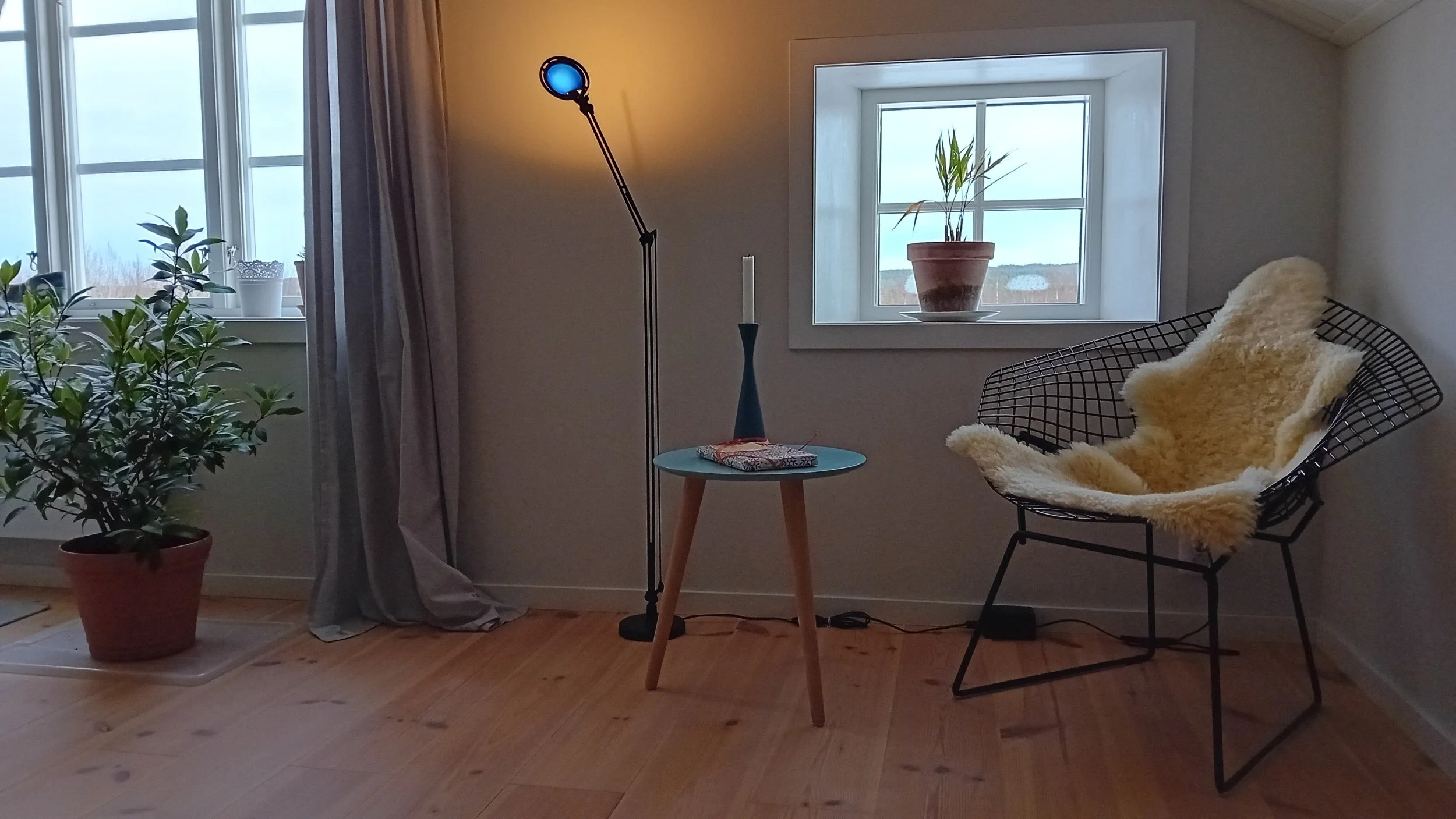 Ein Wohnzimmer mit einem großen Fenster, einem kleinen runden Tisch, einer Stehlampe, einem Korbtisch mit Kerze, einem Sessel mit einem Fell, einem Pflanzenkübel und einigen kleinen Pflanzen auf der Fensterbank.