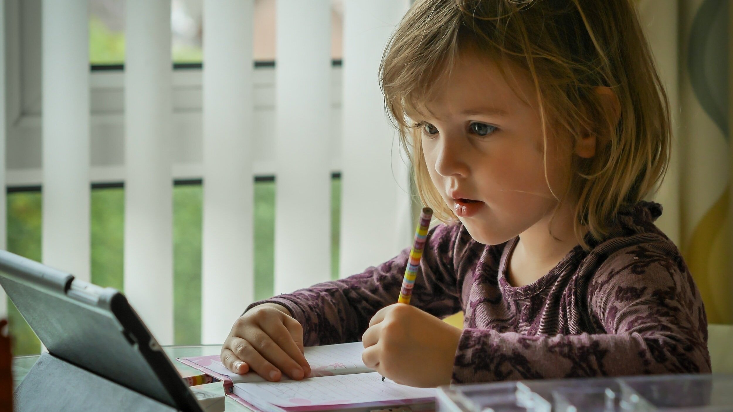A young girl with light brown, wavy hair looking at a tablet screen while holding a rainbow-colored pencil above an open notebook on a table.