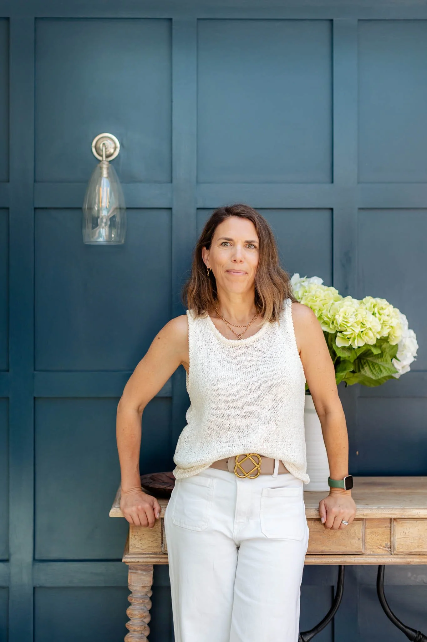 Katie, founder of Hive Social, in a white sleeveless top and pants standing beside a wooden console table with flowers against a dark blue paneled wall