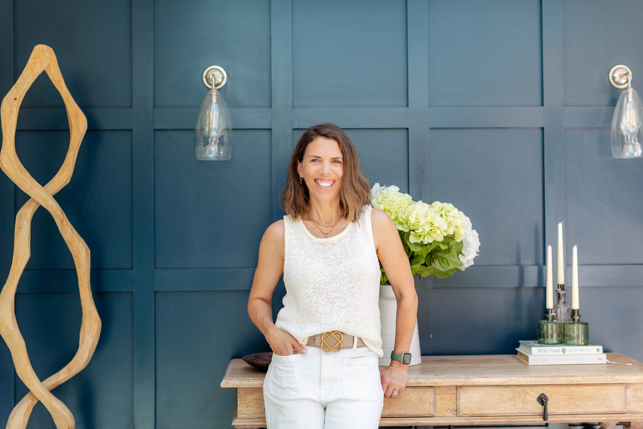 Katie, founder of Hive Social, in a white outfit standing in front of a navy paneled wall with flowers, candles, and wood decor.