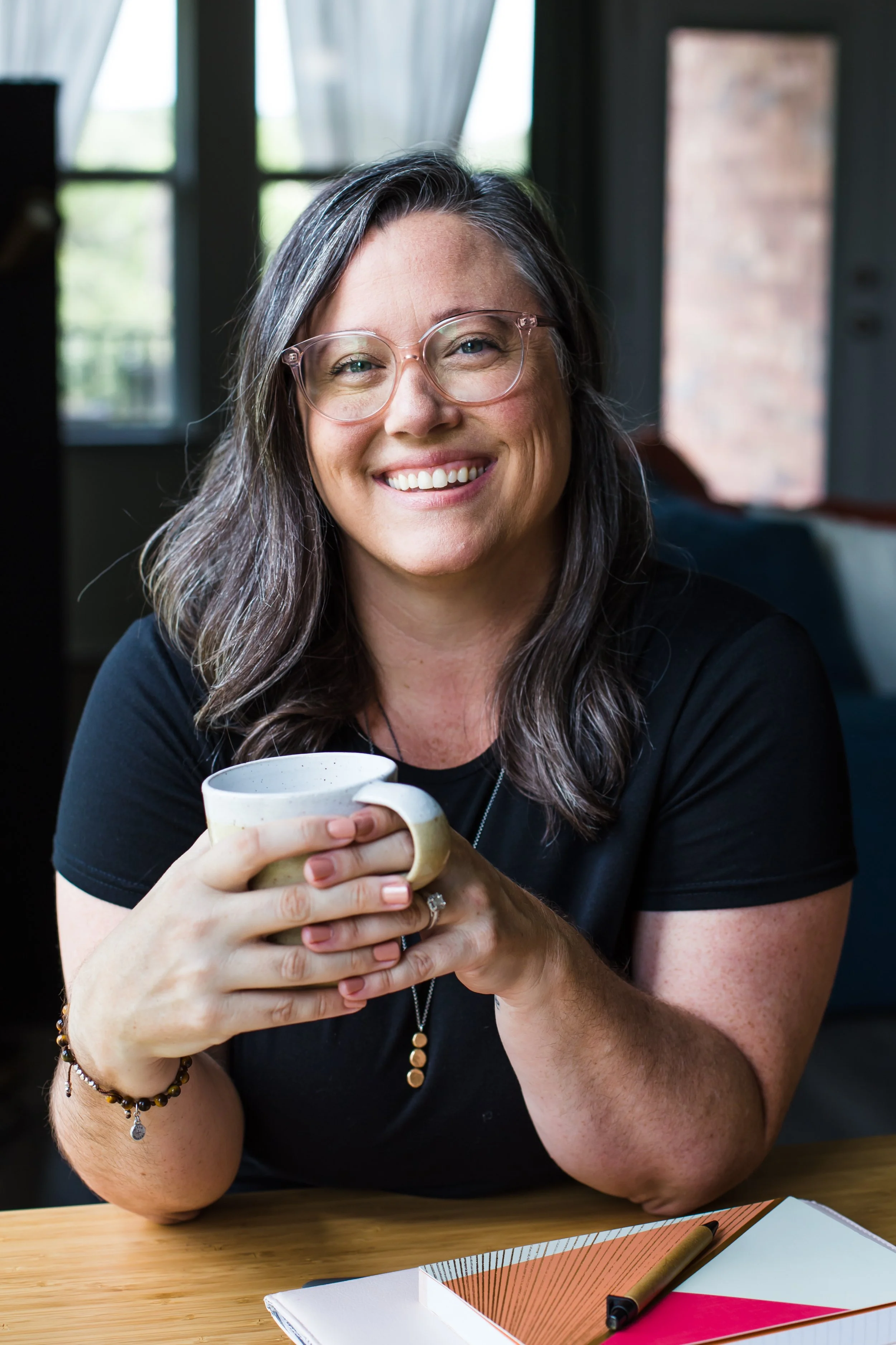 A woman with glasses is smiling and holding a mug with both hands, sitting at a wooden table in a room with large windows and natural light, with a notebook, pen, and some papers on the table.