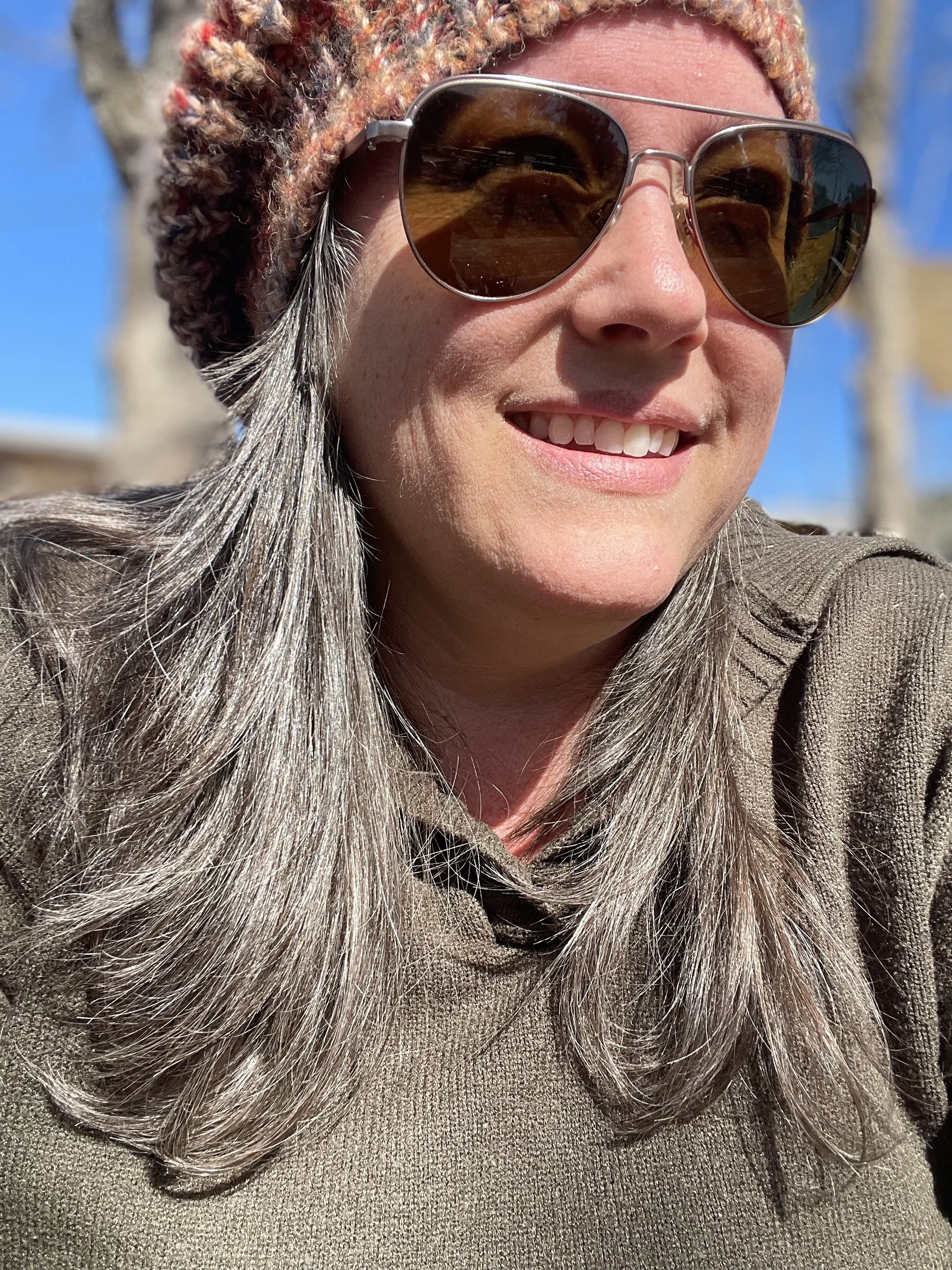 Person outdoors wearing sunglasses, a knitted hat, and a sweater, smiling under sunlight.
