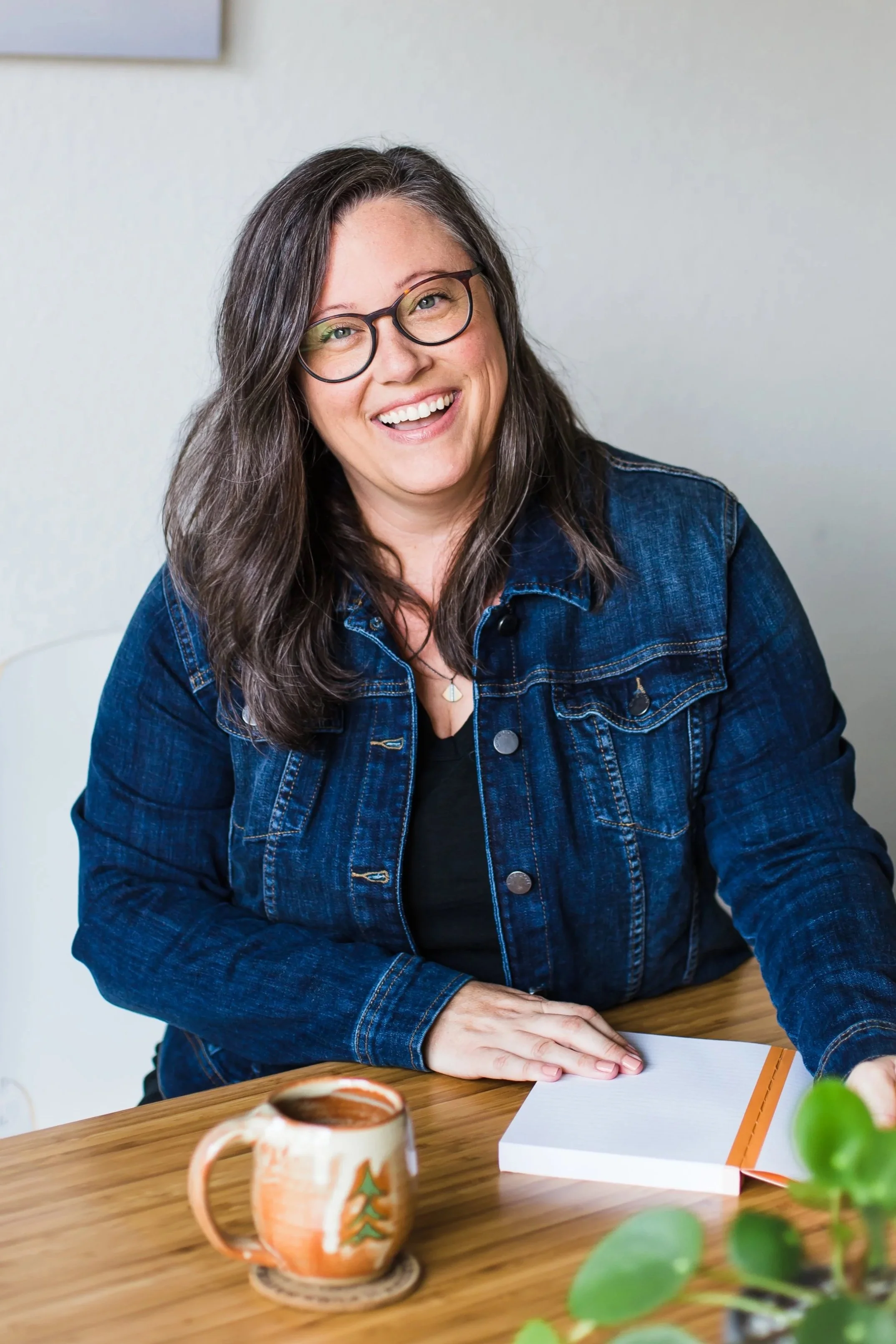 A woman with glasses and long brown hair smiling while sitting at a wooden table. She wears a denim jacket and has a notebook and a ceramic mug with a tree design in front of her.