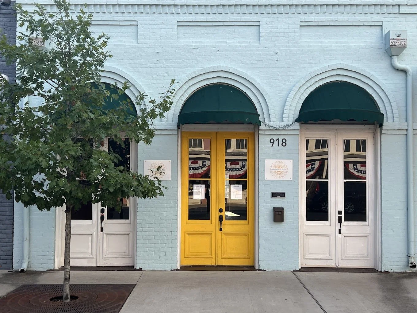 Light blue brick building with three arched awnings and a central yellow door, flanked by two white double doors. A small tree is in front of the left door, and there are patriotic bunting decorations reflected in the glass.