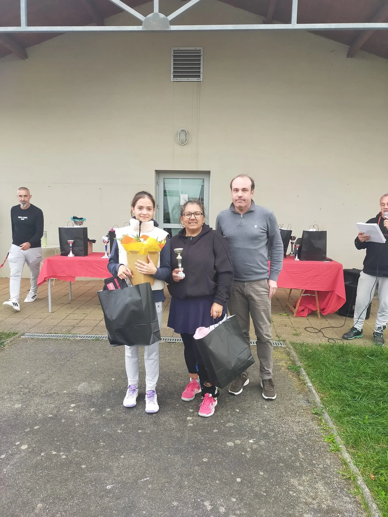 Une cérémonie de remise de prix avec trois personnes au centre, une jeune fille avec un bouquet et un trophée, deux adultes avec des sacs cadeaux devant un tableau de table avec des trophées et des sacs, en extérieur