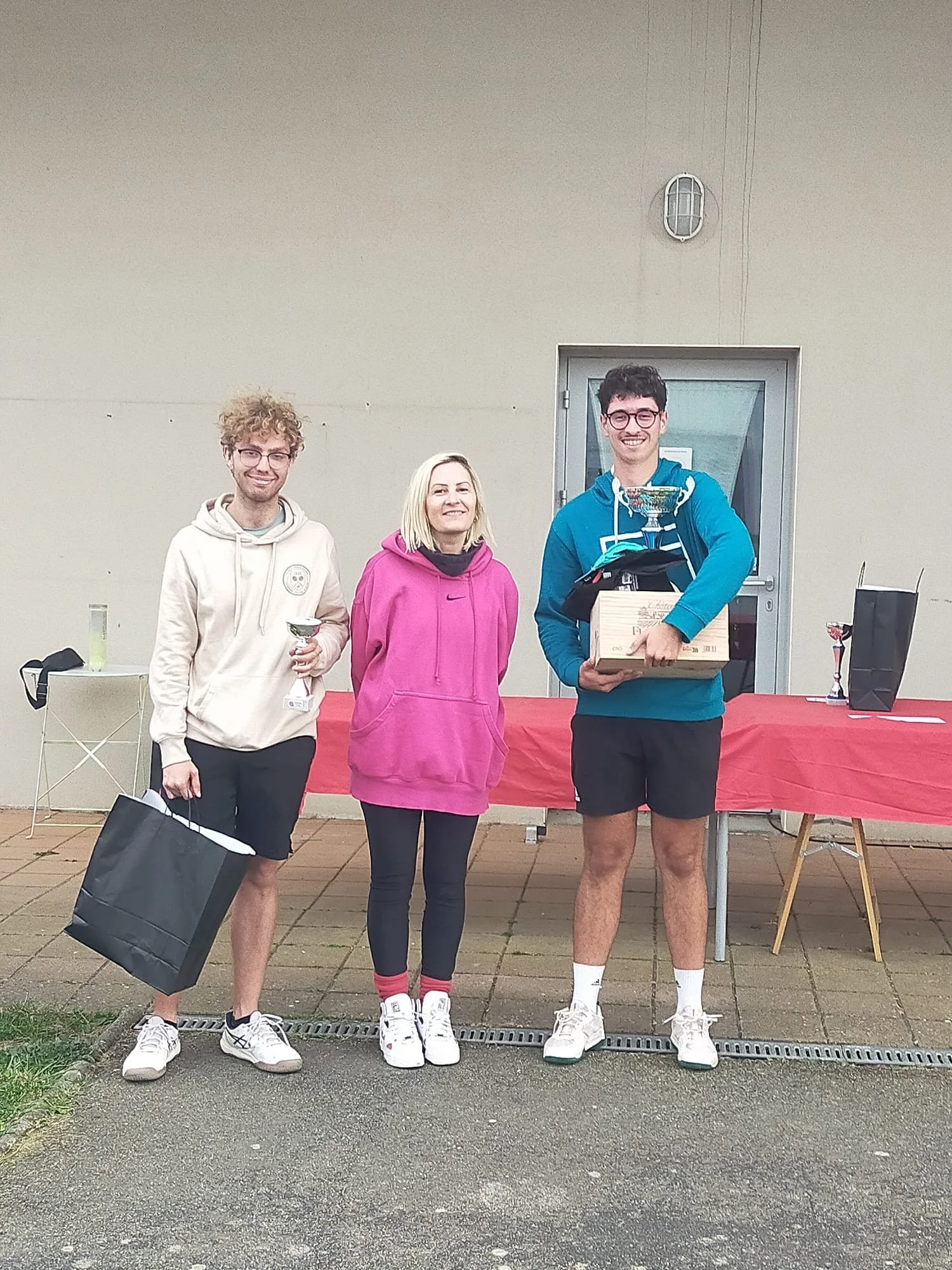 Trois personnes souriantes posent lors d'une cérémonie de remise de prix, deux hommes avec des trophées en main, une femme au centre, en extérieur devant un mur beige, une table avec des trophées et des sacs cadeaux en arrière-plan.