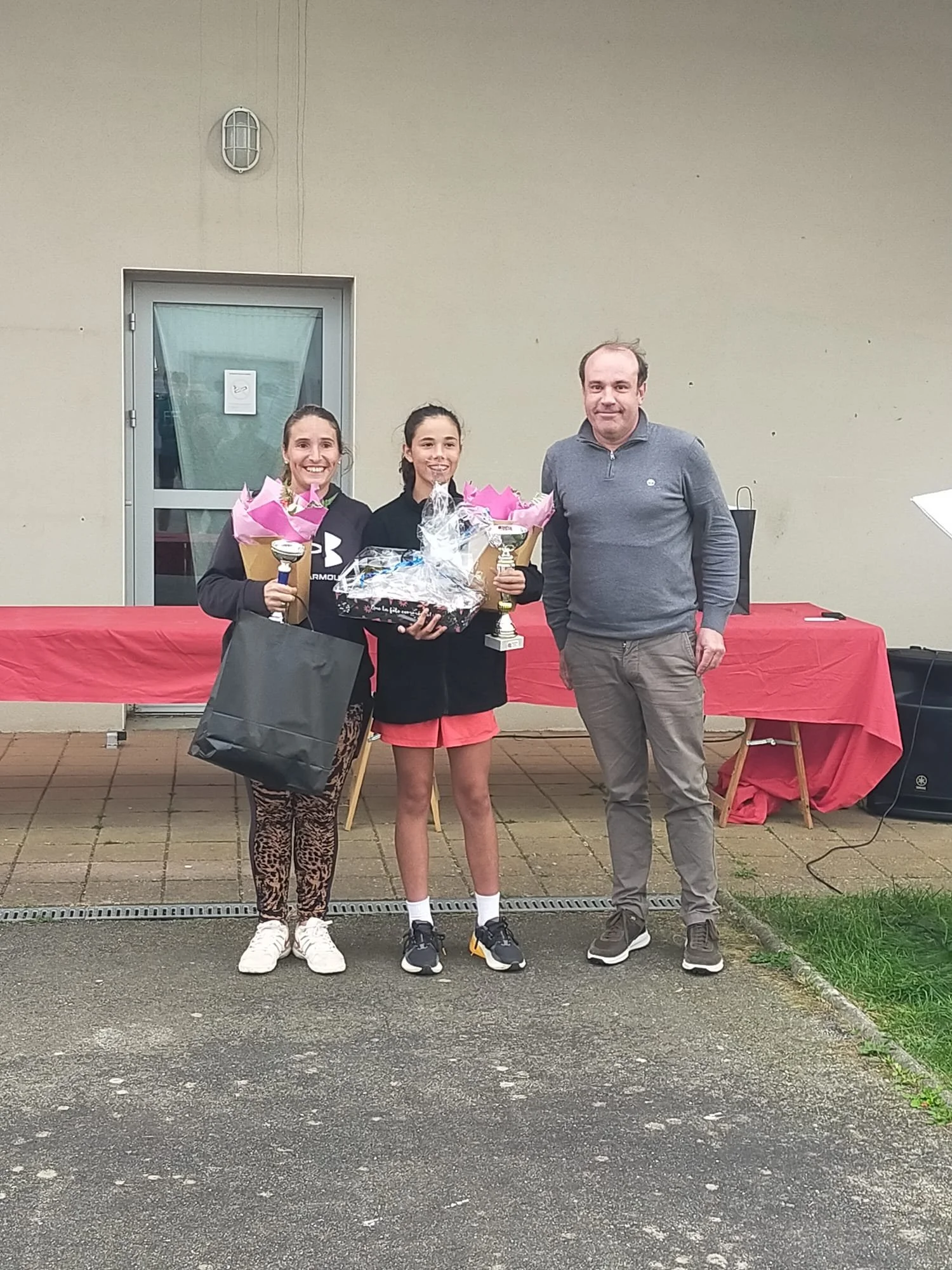 Deux jeunes femmes avec des trophées et bouquets de fleurs, accompagnées d'un homme, lors d'une cérémonie de remise de prix.