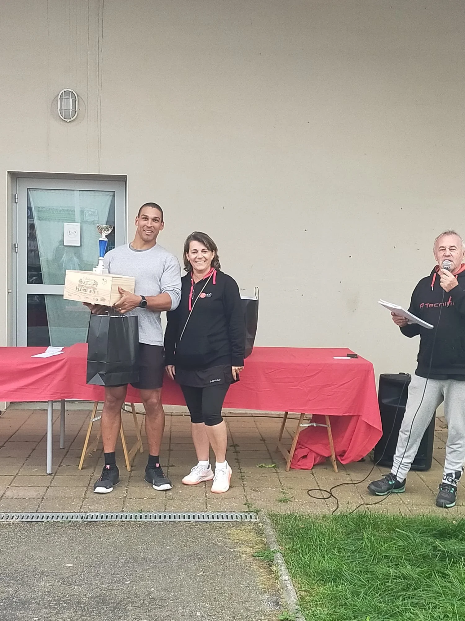Une cérémonie de remise de prix avec un homme tenant un trophée, une femme à côté de lui, et un homme parlant dans un micro, devant une table recouverte d'une nappe rouge, à l'extérieur.