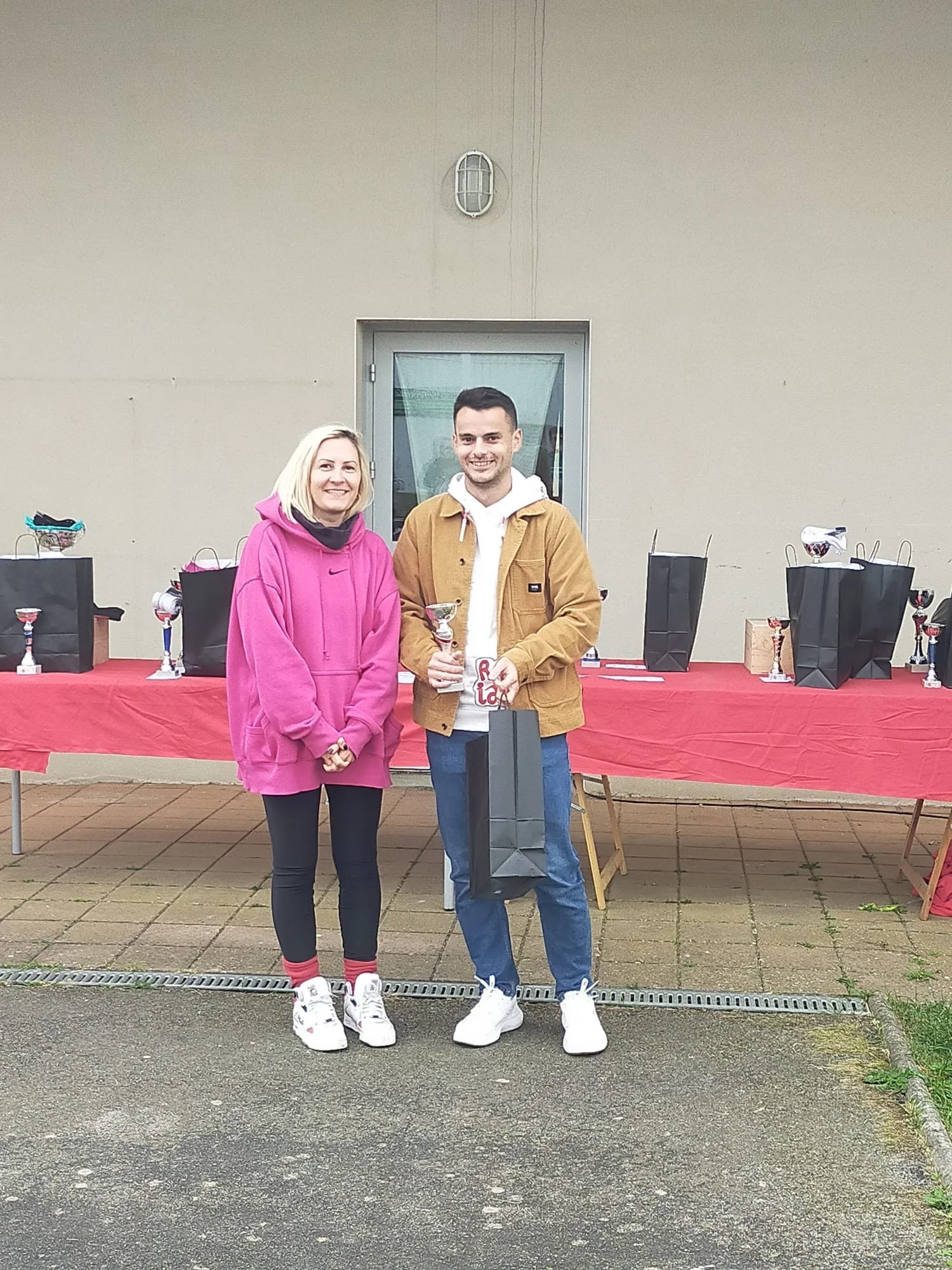 Deux personnes posent avec des trophées lors d'une cérémonie de remise de prix devant une table couverte d'une nappe rouge et plusieurs sacs cadeaux, à l'extérieur d'un bâtiment.