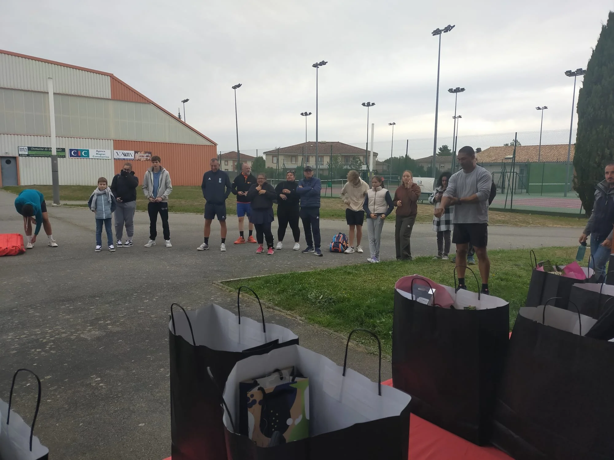 Groupe de personnes, enfants et adultes, participant à une activité en extérieur près d'un terrain de tennis, avec des sacs cadeaux ou sacs de rangement à l'avant.