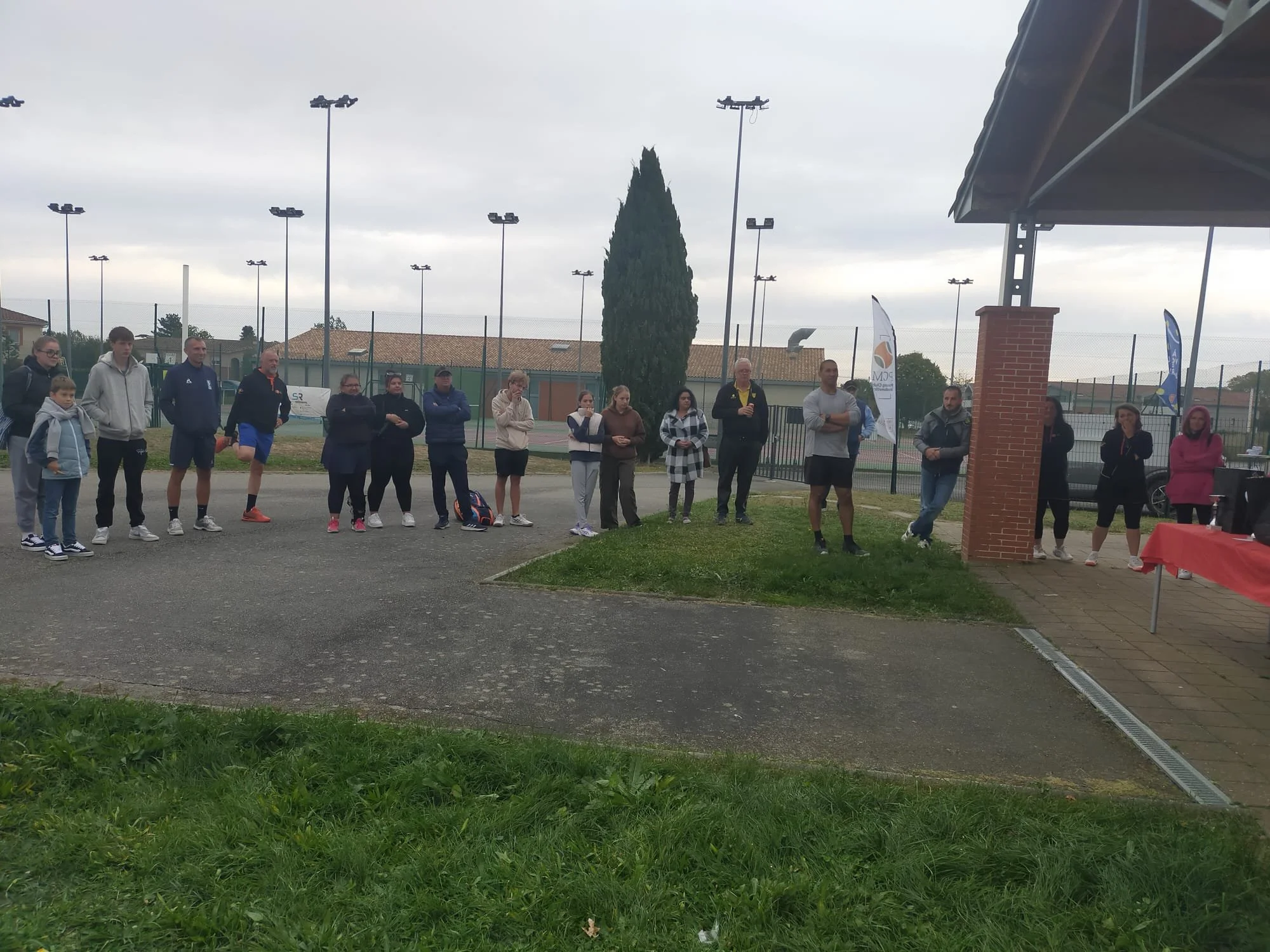 Groupe de personnes debout en ligne à l'extérieur près d'une tente, probablement lors d'un événement ou d'une activité sportive, avec un terrain de tennis en arrière-plan et des drapeaux.