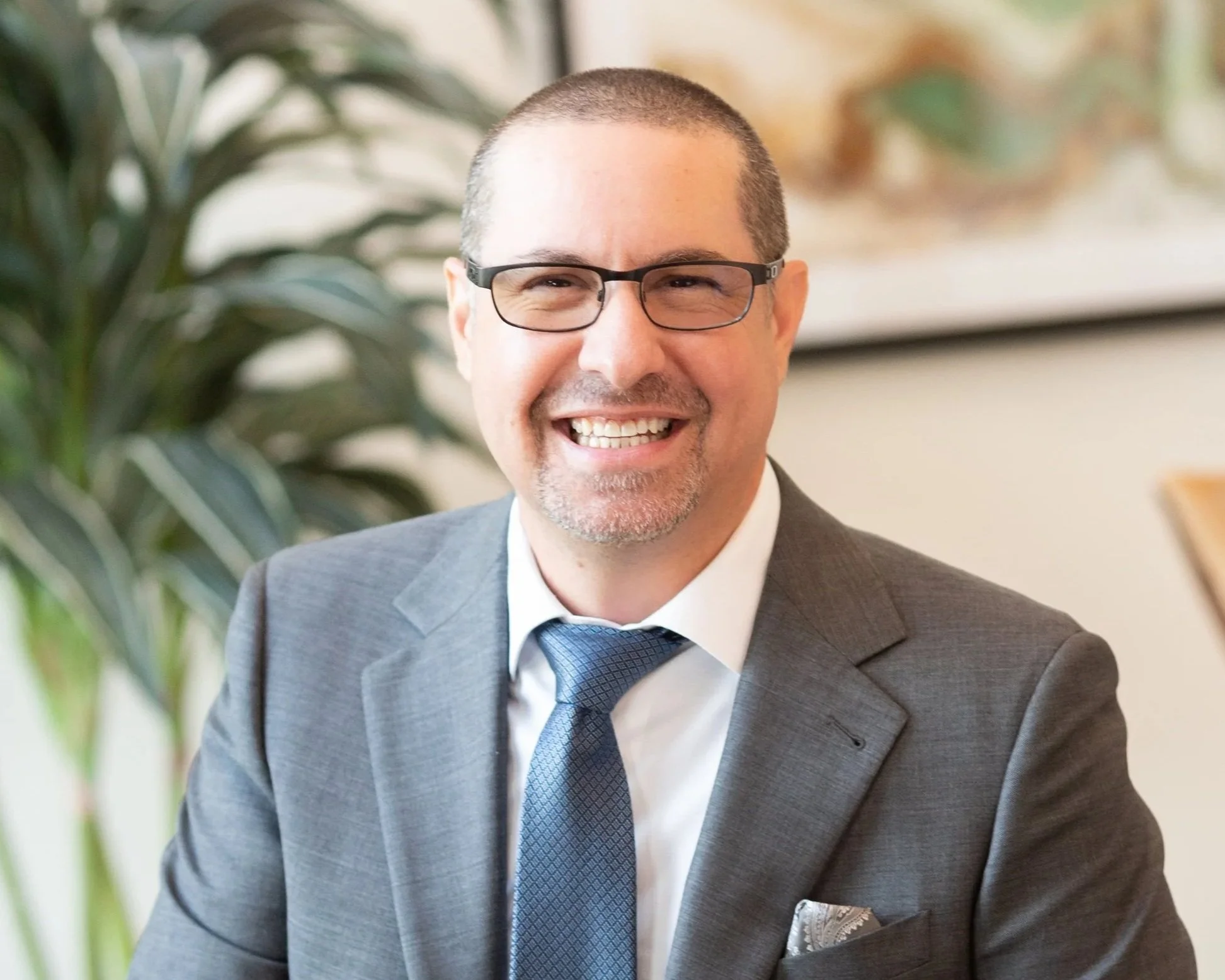A smiling man wearing glasses, a gray suit, white shirt, and blue tie sitting in an office with a plant and abstract art on the wall.