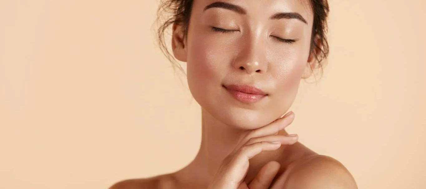 Close-up of a woman with clear, glowing skin gently touching her chin with her hand, eyes closed, against a neutral beige background.