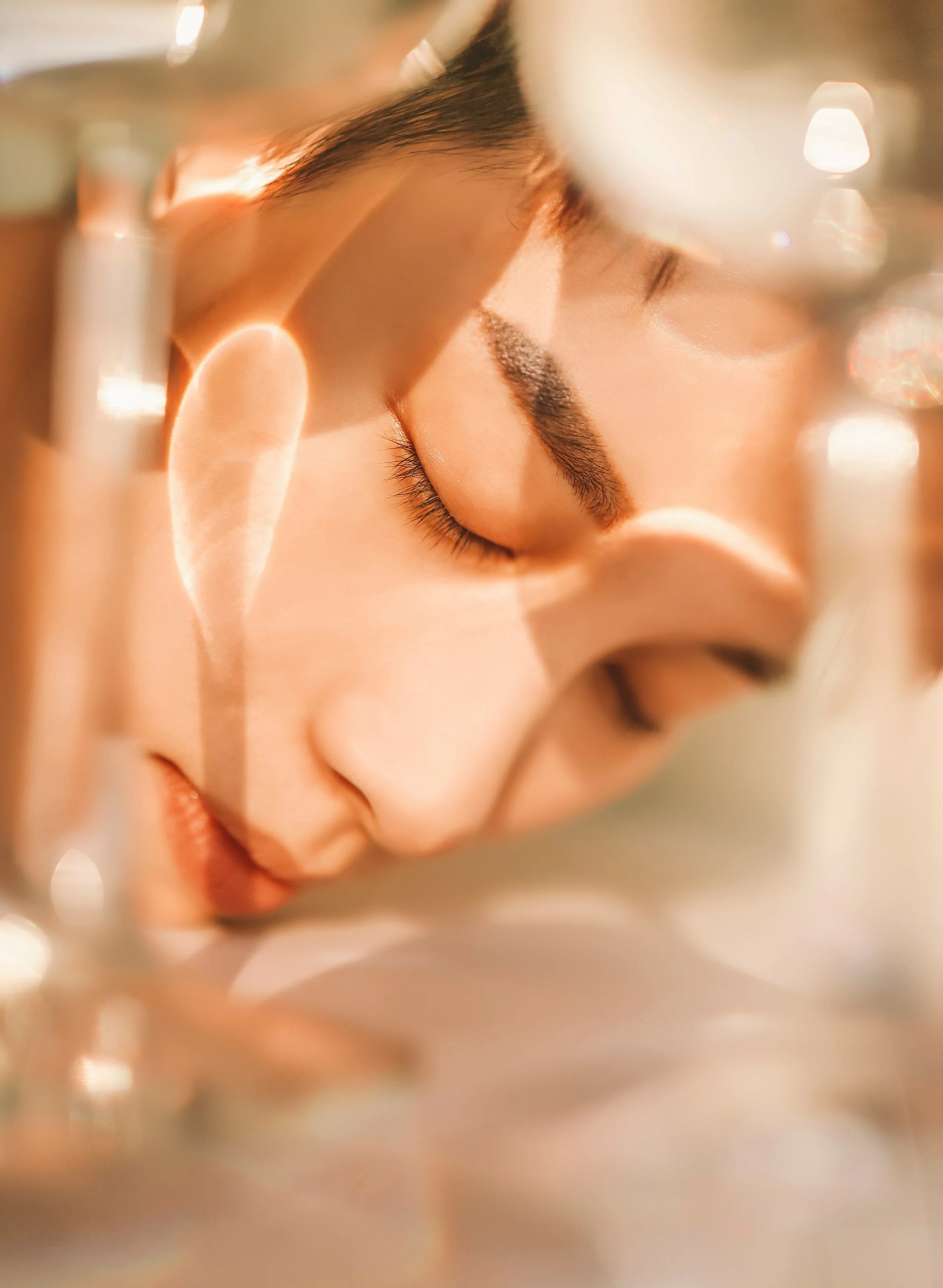 Close-up of a woman's face with eyes closed, seen through a glass, creating a blurred effect with warm lighting.