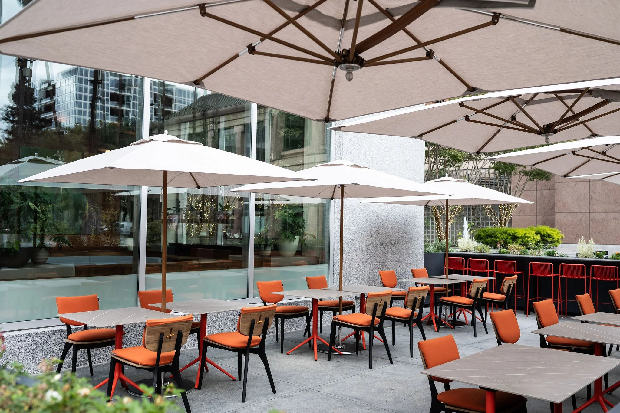 An outdoor restaurant patio featuring grey tables and orange chairs under large white umbrellas, highlighting a sun-drenched California dining atmosphere.   Furniture Detail: A detail shot of cognac leather restaurant chairs and textured stone