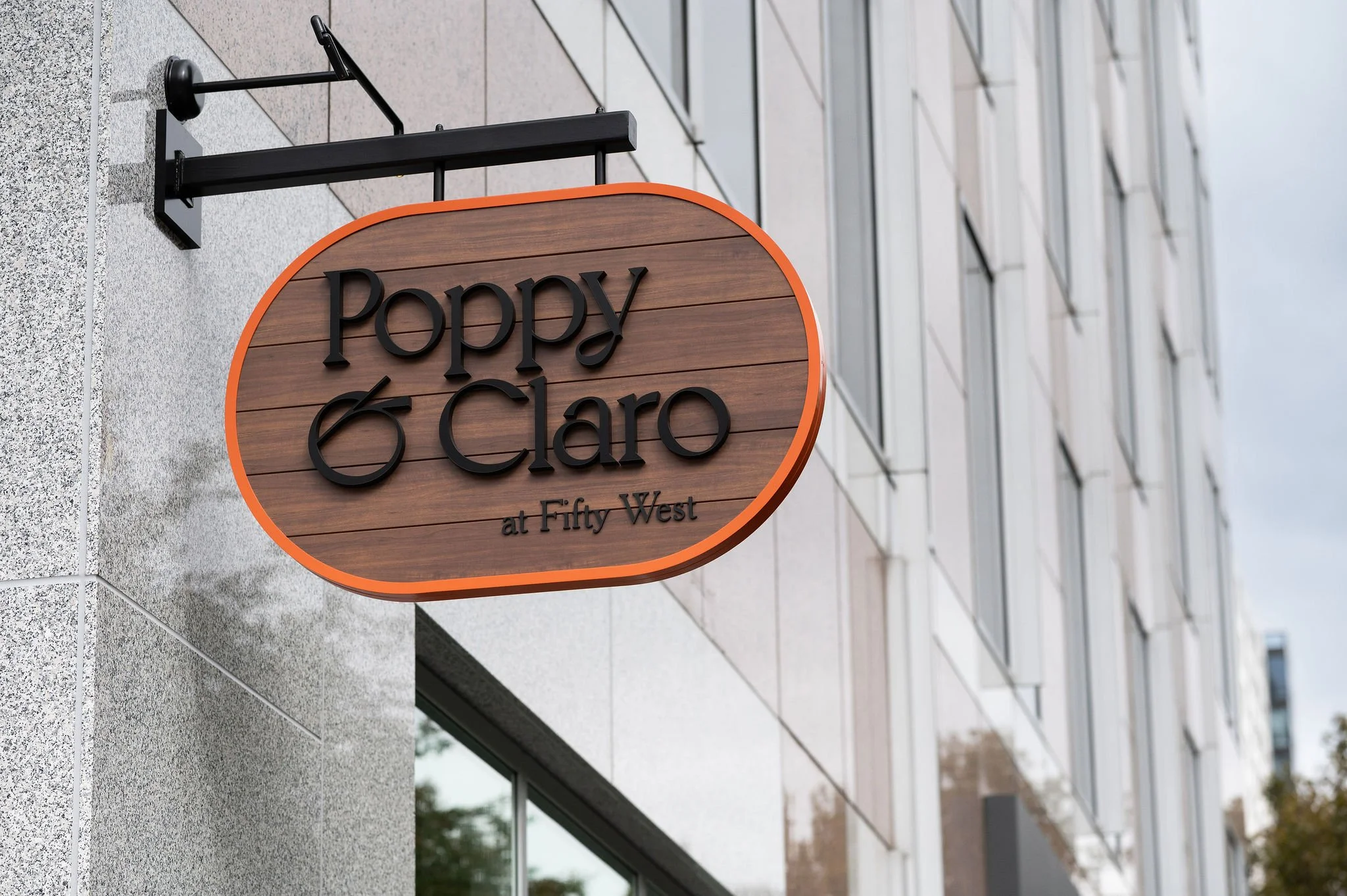 An oval wooden exterior sign for Poppy & Claro at Fifty West with black 3D lettering and a bright orange border, mounted on a light stone building.