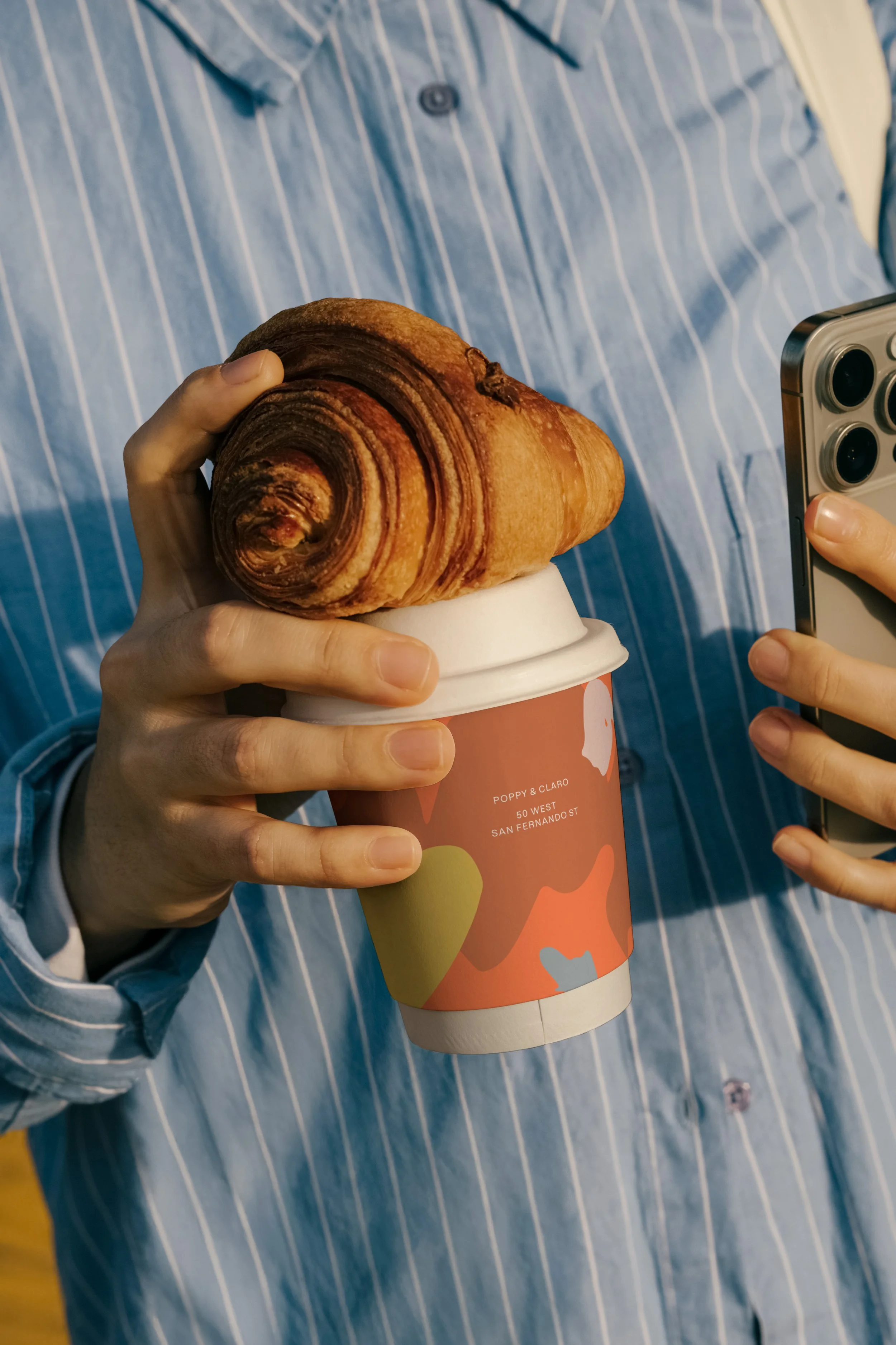 A lifestyle photograph of a guest holding a branded coffee cup with a flaky croissant balanced on top, ideal for a restaurant website design focusing on the "sunlit ease" of morning cafe service.