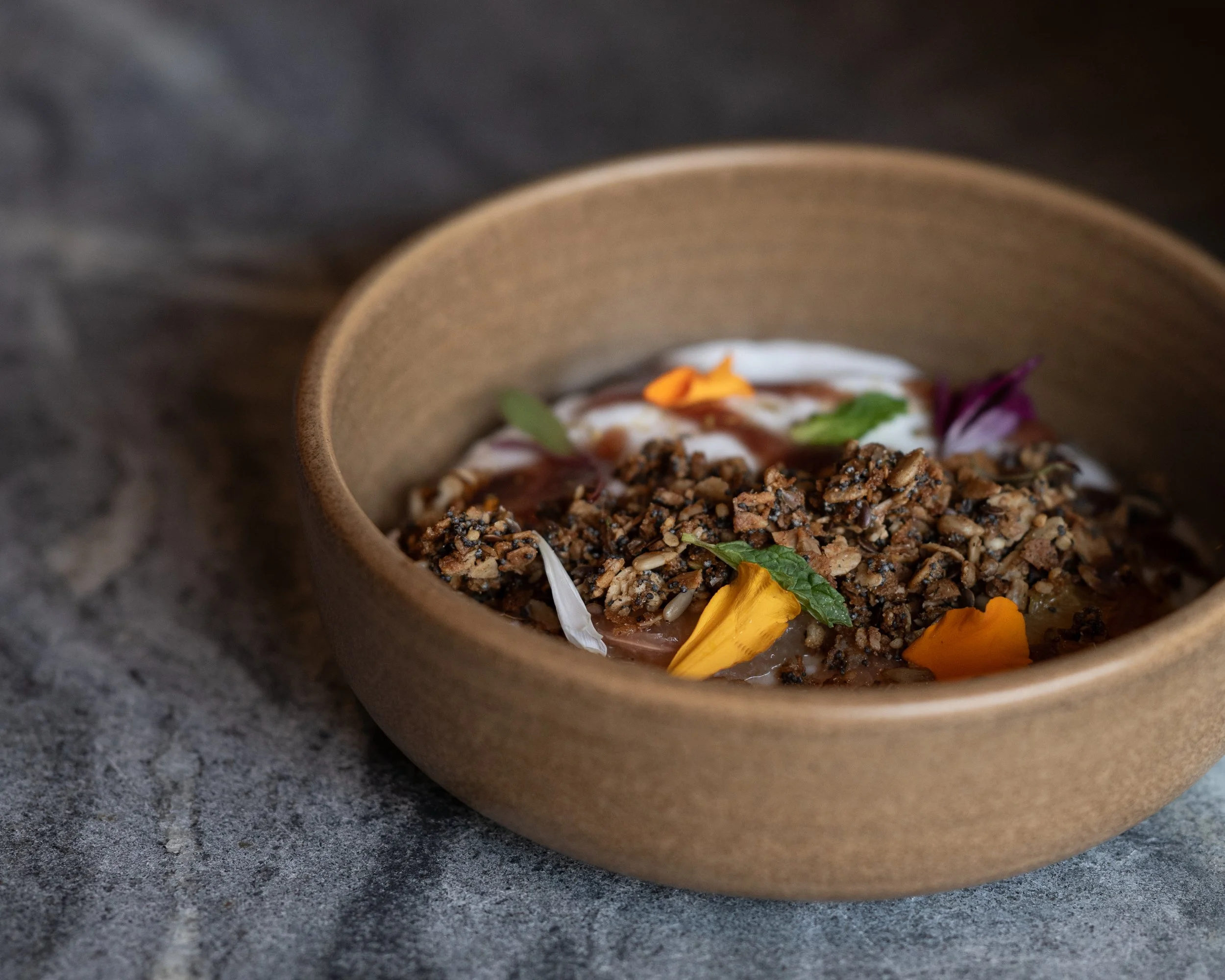 A close-up, professional food photograph of a seasonal breakfast bowl containing dark grains, cream, and orange edible flower petals, reflecting a "globally inspired" menu.