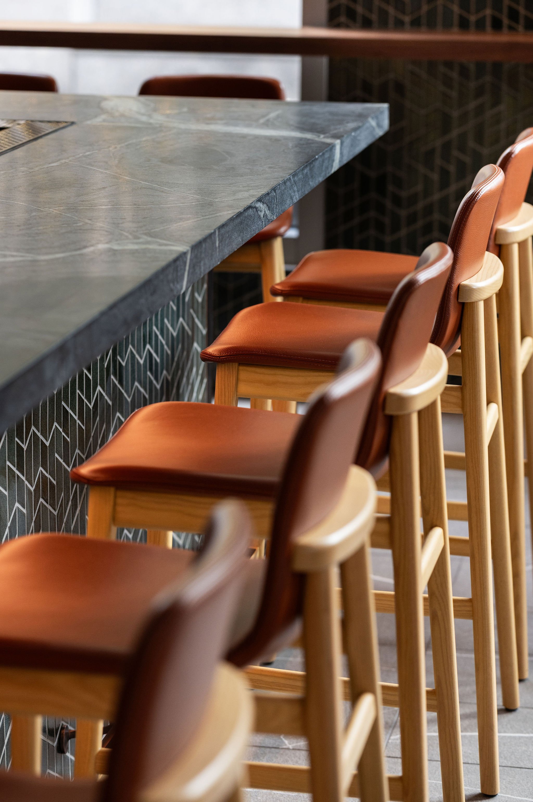 A detail shot of cognac leather restaurant chairs and textured stone countertops, emphasizing the "elevated yet approachable" interior design.