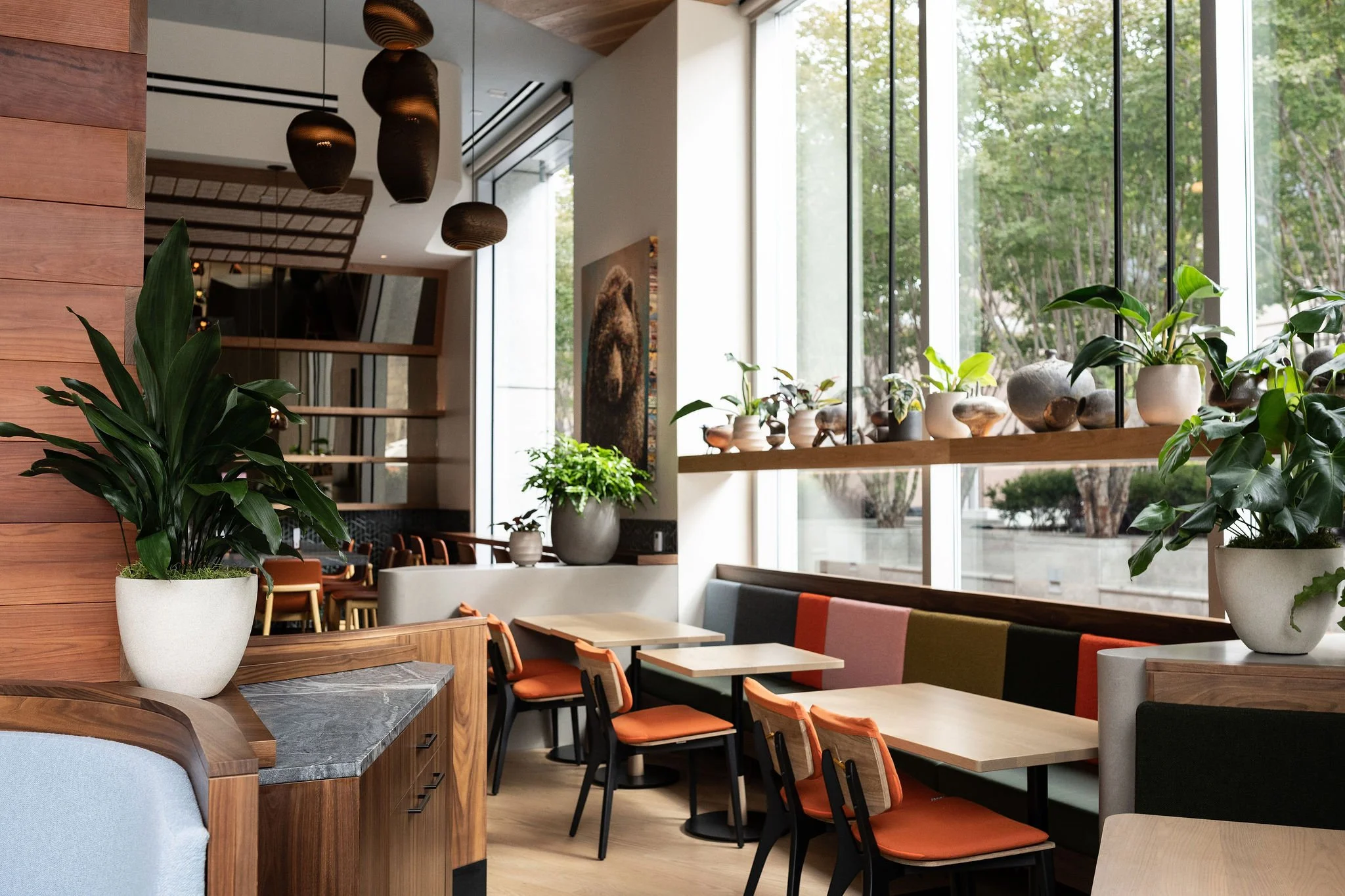 A high-resolution interior photograph used in restaurant website design to showcase a bright, airy dining space with floor-to-ceiling windows, mid-century modern furniture, orange chairs, and decorative potted plants.