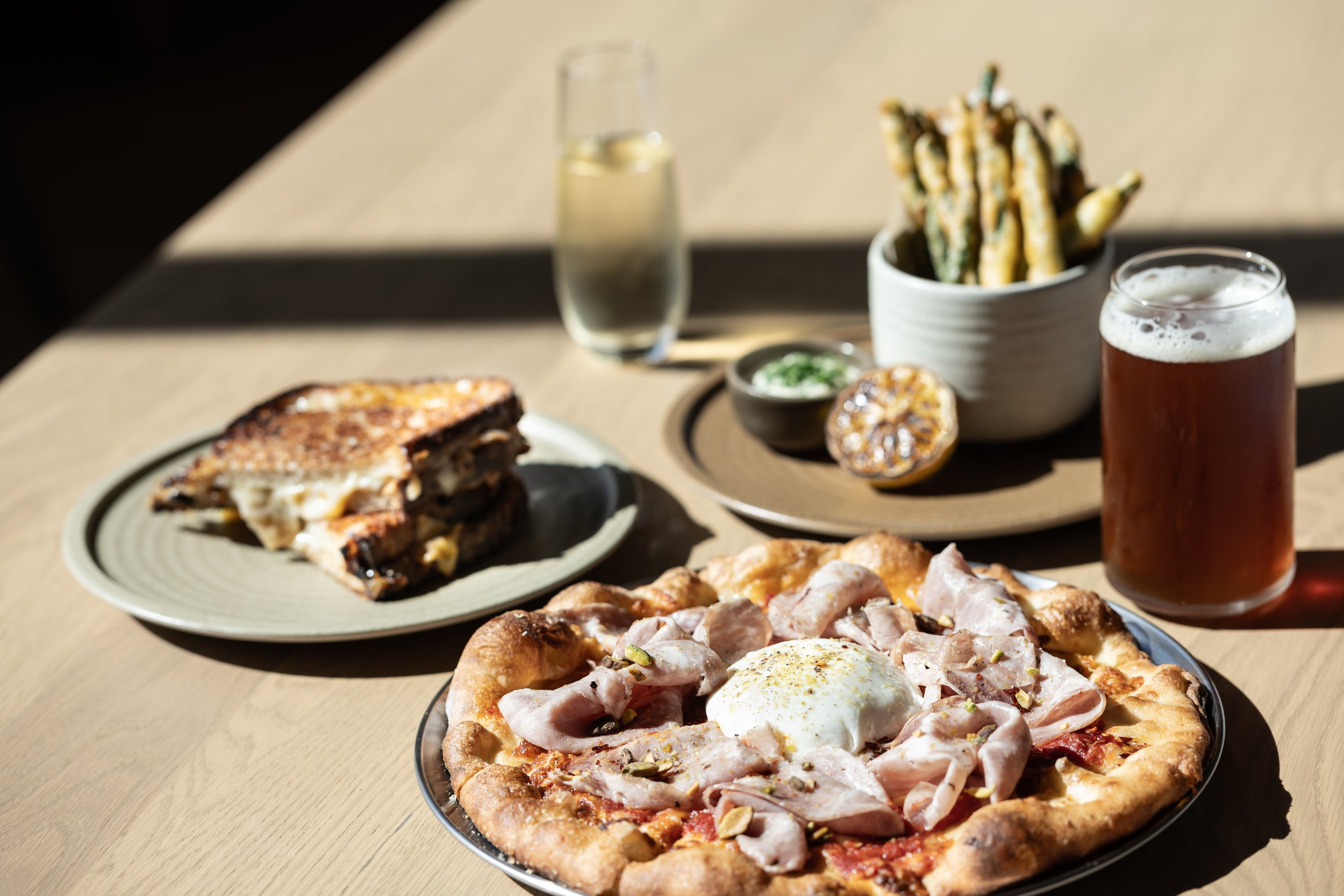 A sunlit table setting featuring a gourmet pizza topped with burrata and mortadella, alongside a toasted panini, intended for a restaurant website design gallery.