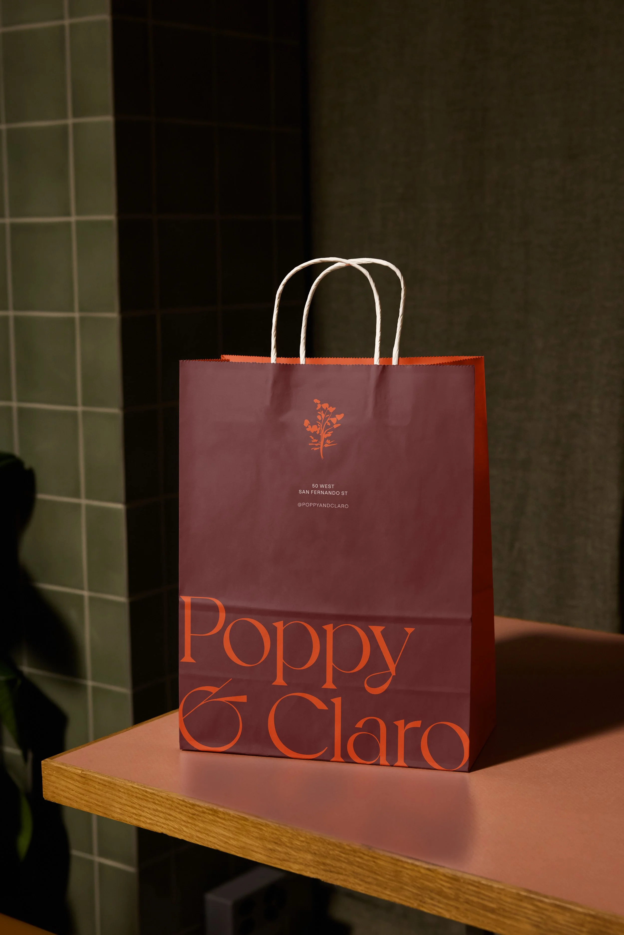 A premium burgundy paper takeout bag featuring the Poppy & Claro logo in vibrant orange, showcased on a restaurant website design to highlight a cohesive and elevated brand identity.