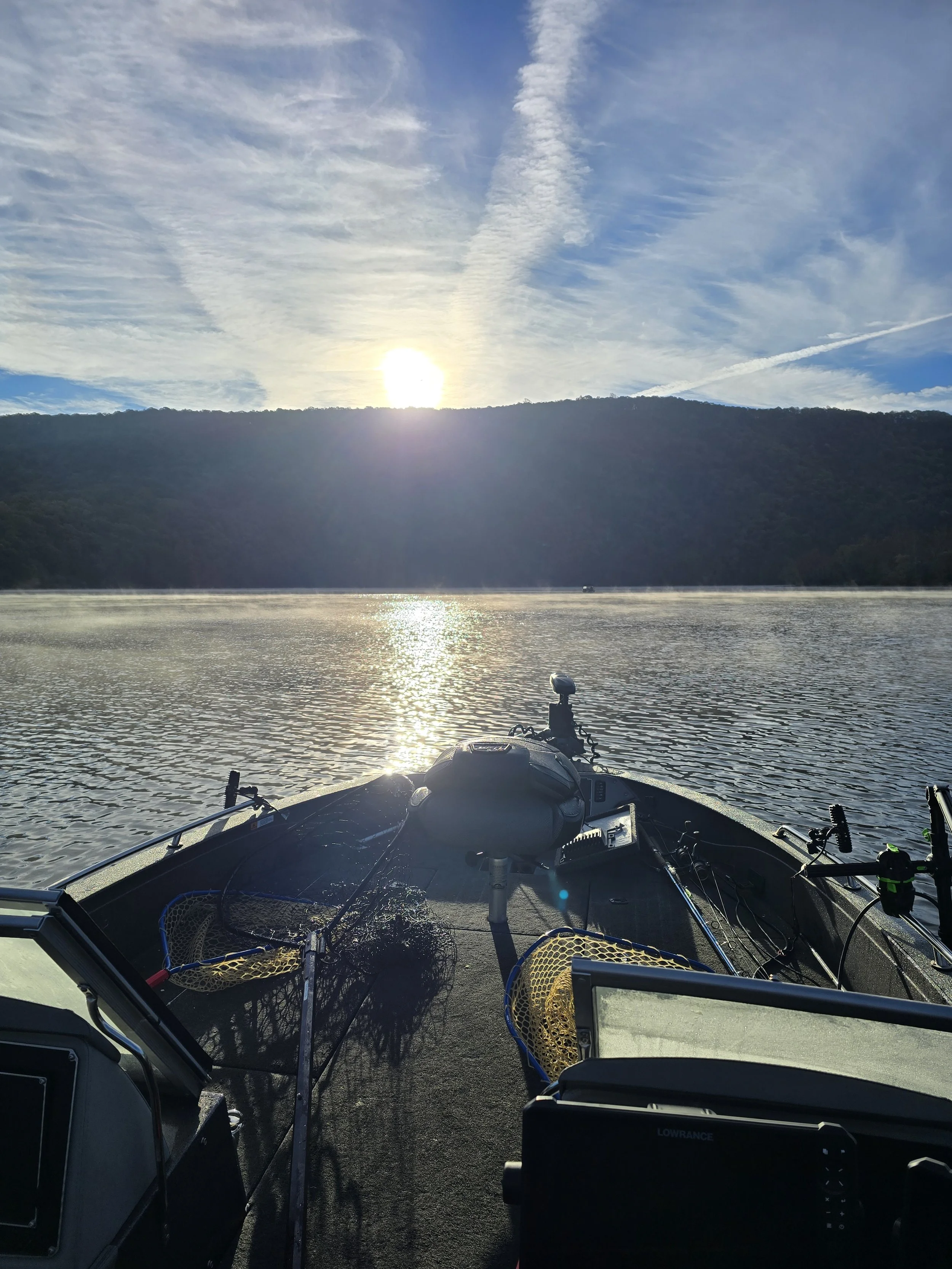 View from a boat on a lake at sunset, with fishing equipment and a fish finder visible, distant hills and a sky with clouds and the sun setting.
