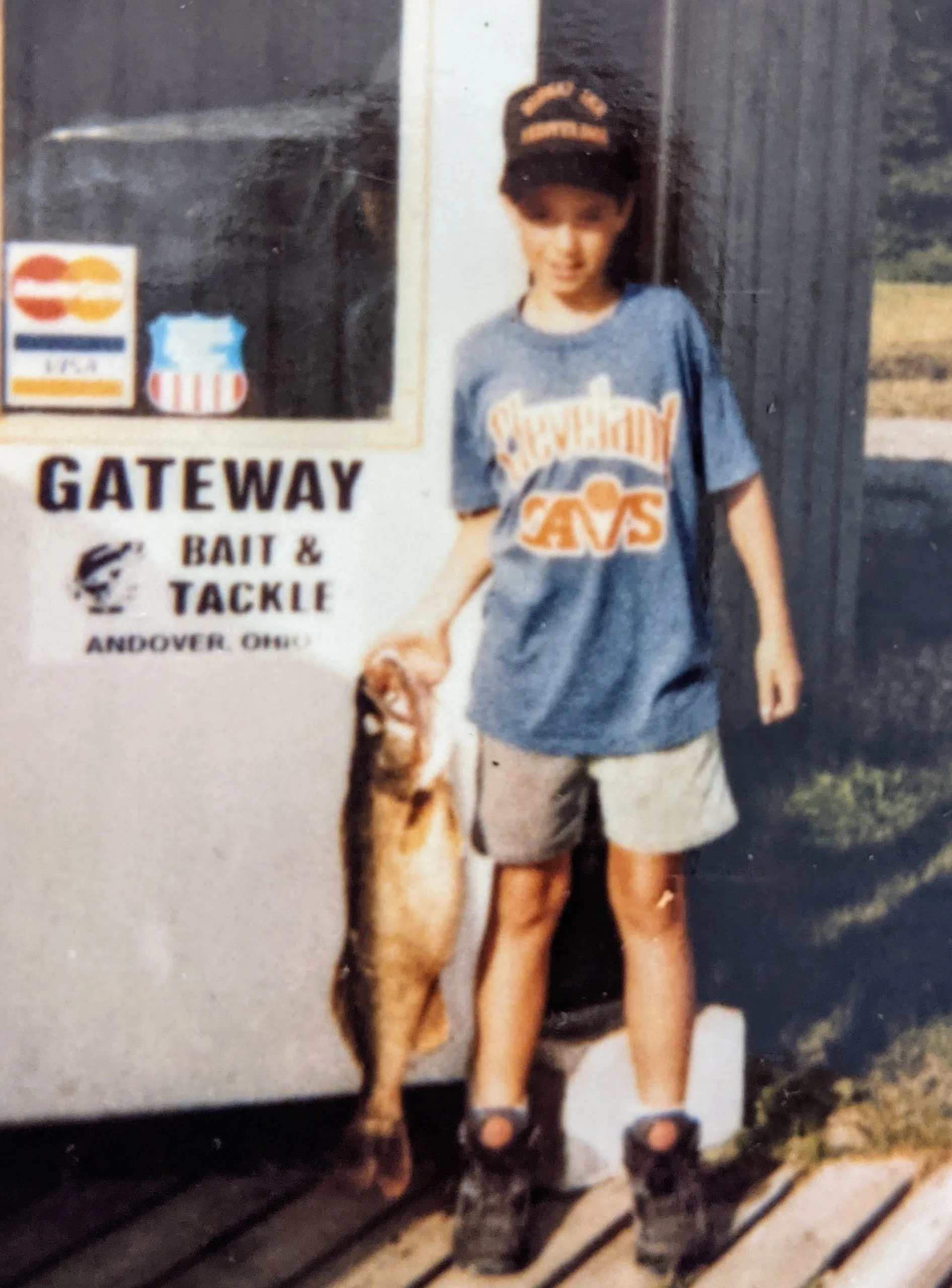 A young boy wearing a Cavs shirt, shorts, Reebok pumps, and a ballcap with Midway Inn his grandparents restaurant, holding a fish Ohio walleye outdoors near a sign that says 'Gateway Bait & Tackle' in Andover, Ohio.