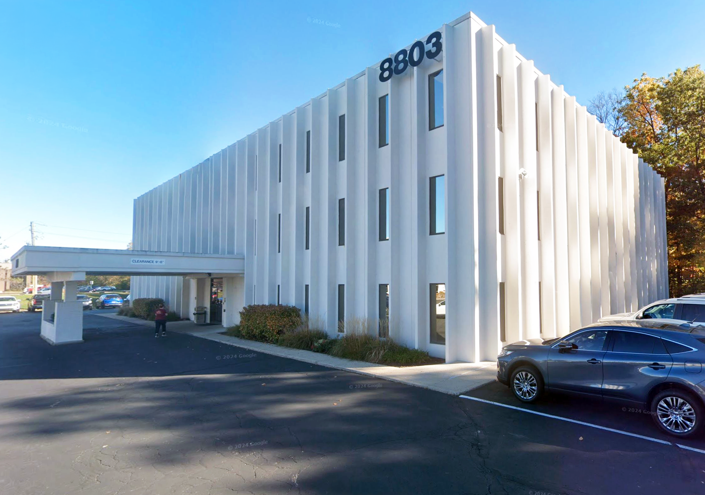 Modern white multi-story building with the number 8803 displayed on the top right corner and a parking lot in front with parked cars and some people walking.