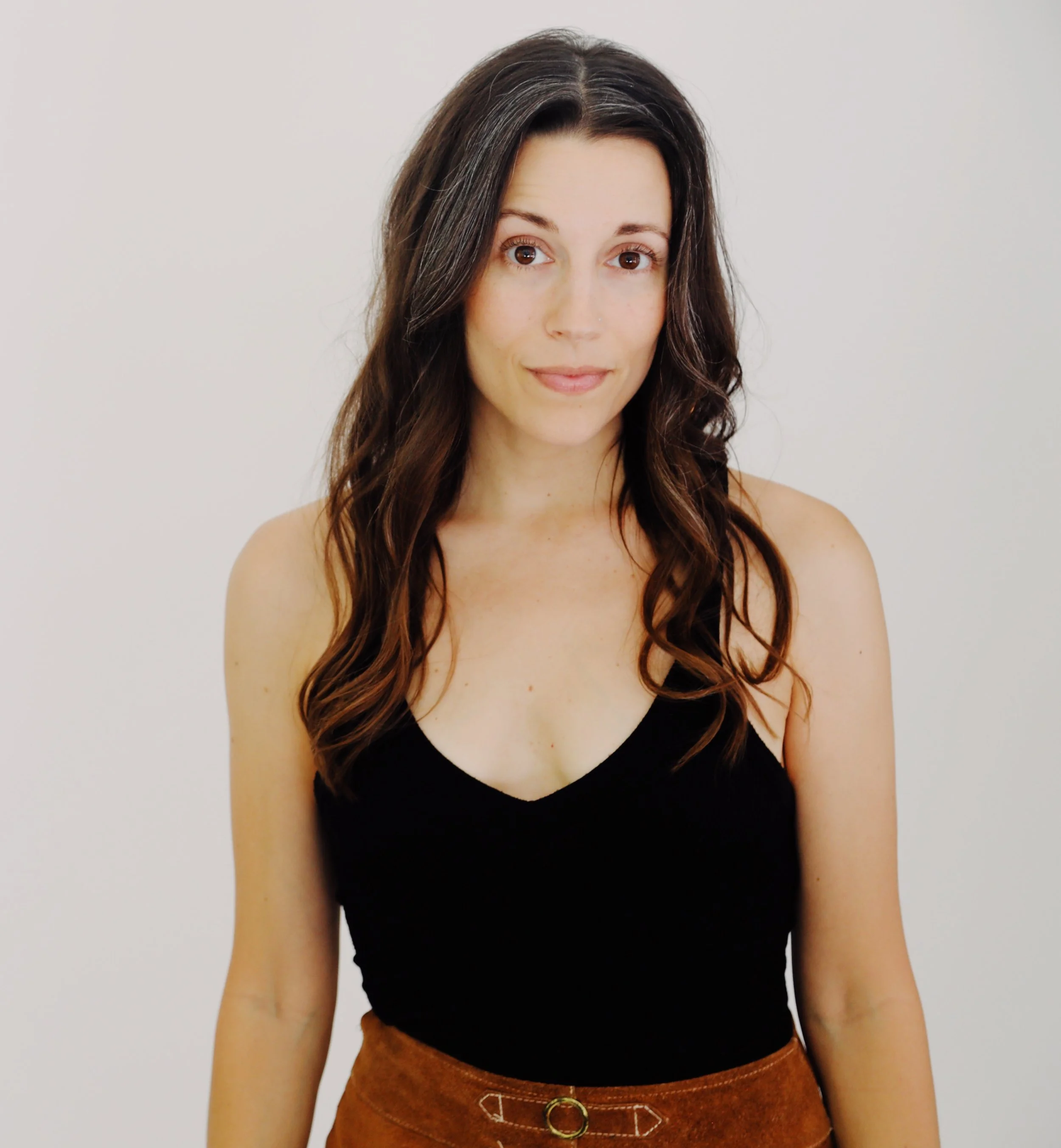 A woman with long brown wavy hair, wearing a black sleeveless top and a brown skirt, standing against a plain white background.
