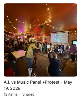 People gathered inside a dimly lit venue with string lights, a large projection screen, and a music panel event titled 'A.I. vs Music Panel +Protest.' Some individuals are filming, and others are seated, watching the presentation.