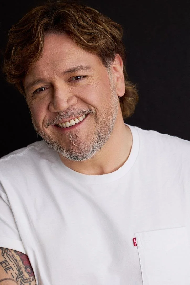 A smiling man with wavy brown hair and a beard, wearing a white T-shirt, against a black background.
