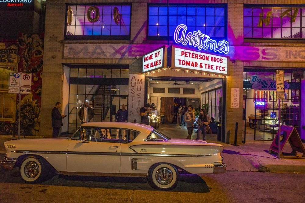 A vintage cream-colored car parked in front of the Antones music venue in downtown Austin. The venue has bright neon signs and colorful lighting, with several people gathered outside.